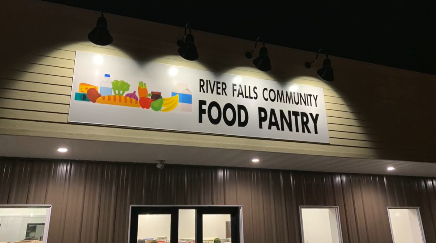 Sign reading 'River Falls Community Food Pantry' with food illustrations, illuminated at night.