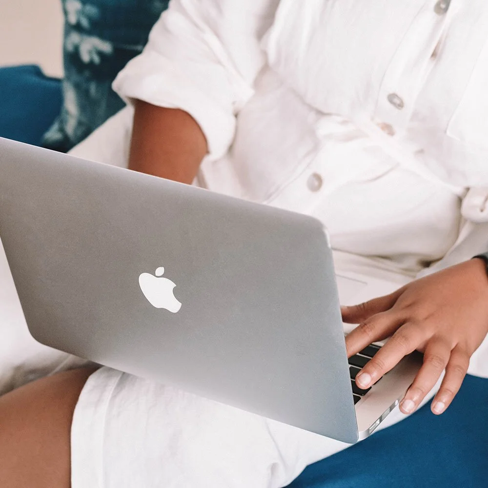 Person using a silver MacBook laptop, sitting on a blue couch, wearing a white shirt and white shorts.