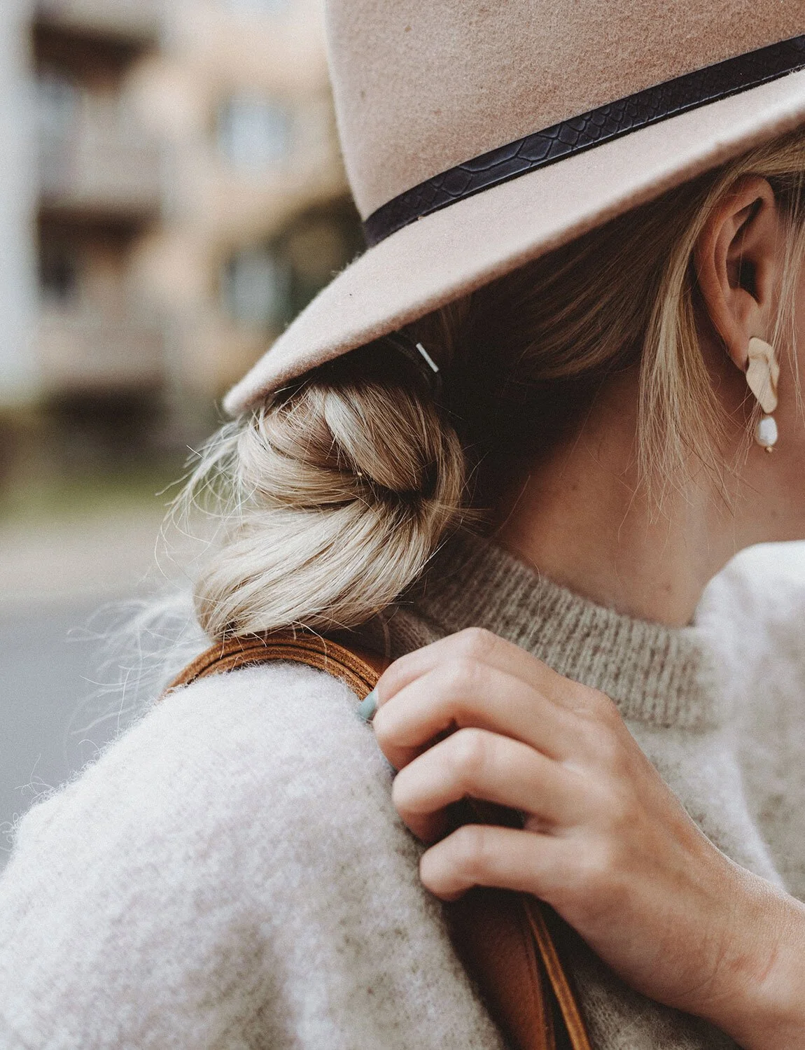 Close-up of a woman wearing a beige hat with a black band, a cozy beige sweater, and dangling earrings, holding a brown leather strap over her shoulder.