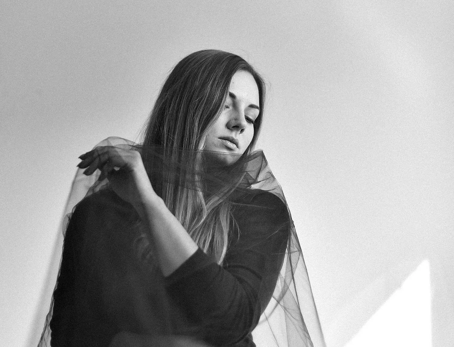 A black and white portrait of a woman with long hair, eyes closed, wearing a sheer fabric covering her shoulder.
