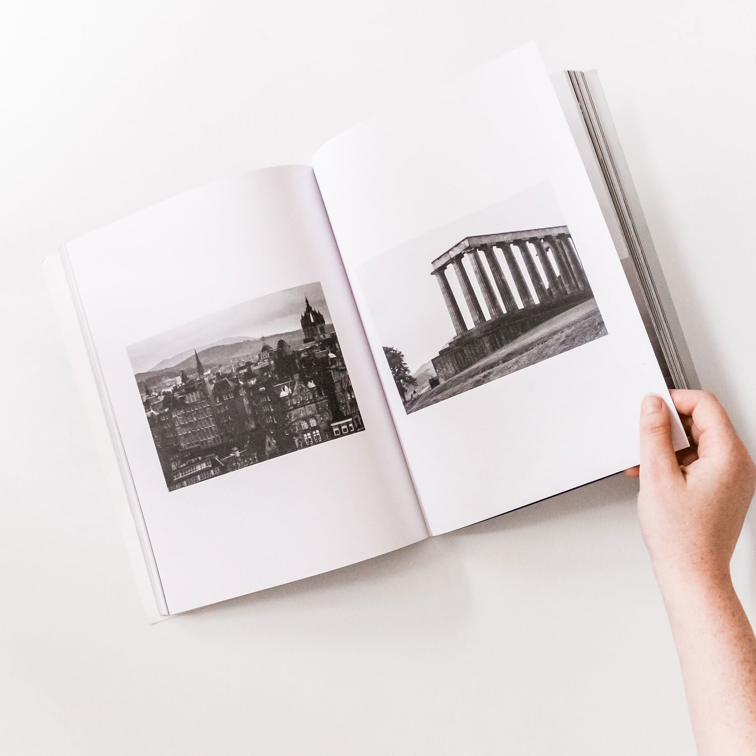 An open book with black-and-white photographs of cityscape and classical building with columns, held by a person's hand on a white surface.