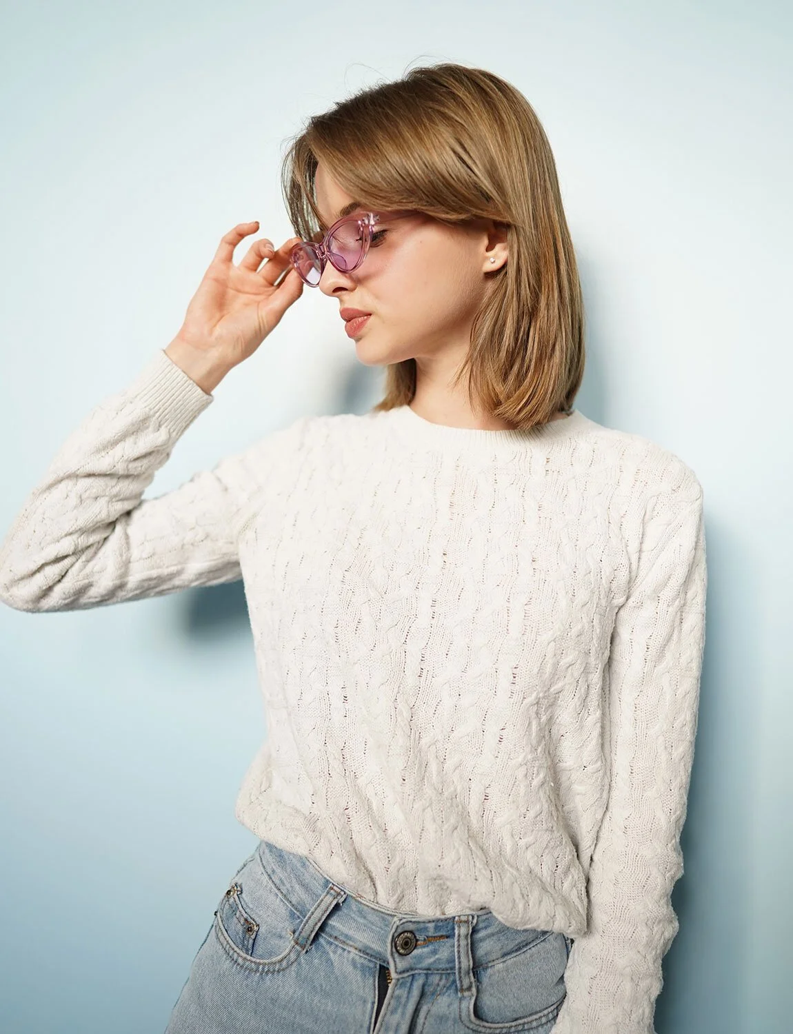 Young woman with shoulder-length brown hair and pink sunglasses, wearing a cream cable-knit sweater and light blue jeans, standing against a light blue background, looking down and adjusting her sunglasses.