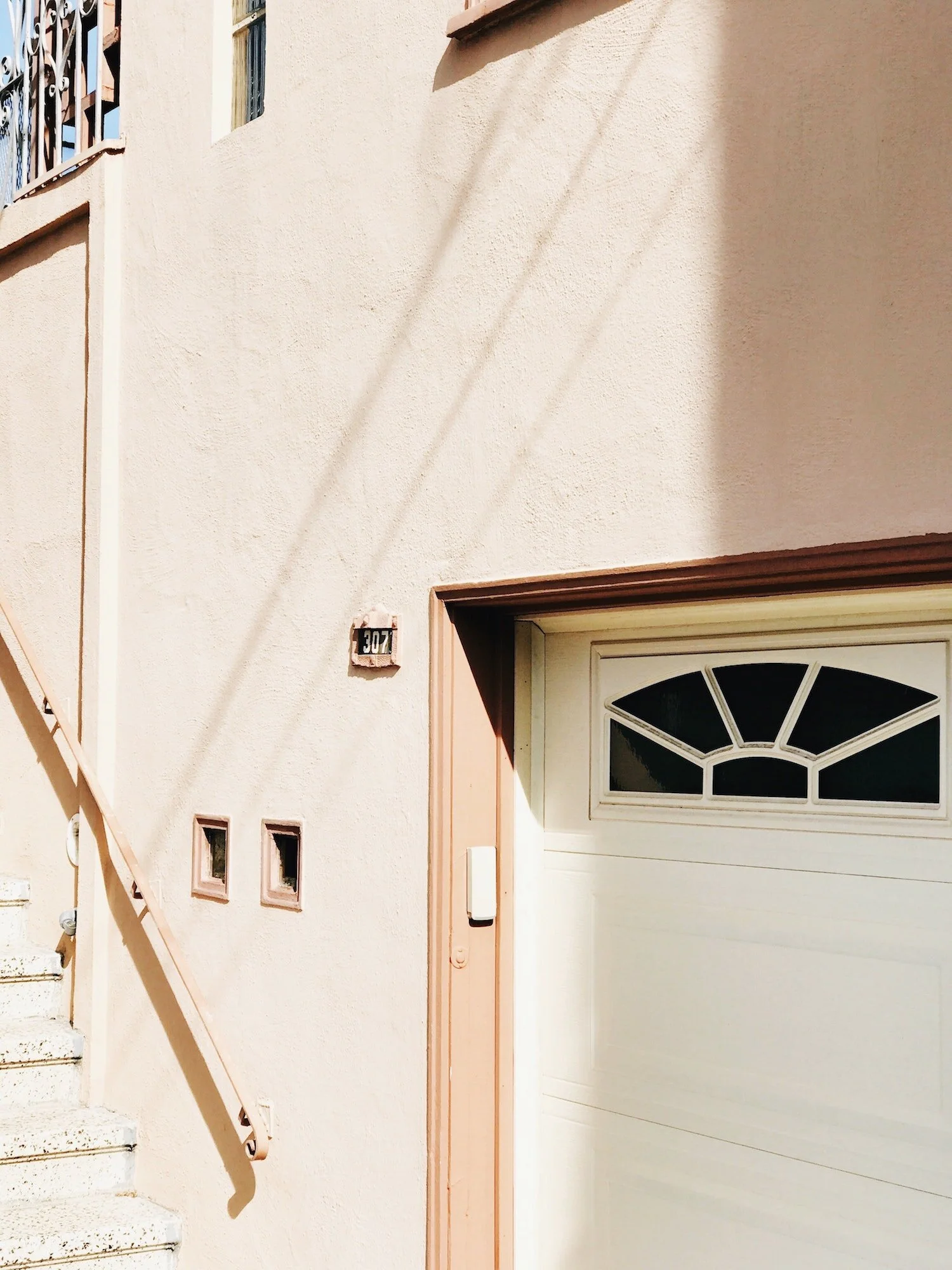 A beige stucco house exterior showing a garage door with a decorative window, a staircase with a metal railing, and a house number 307 on the wall.