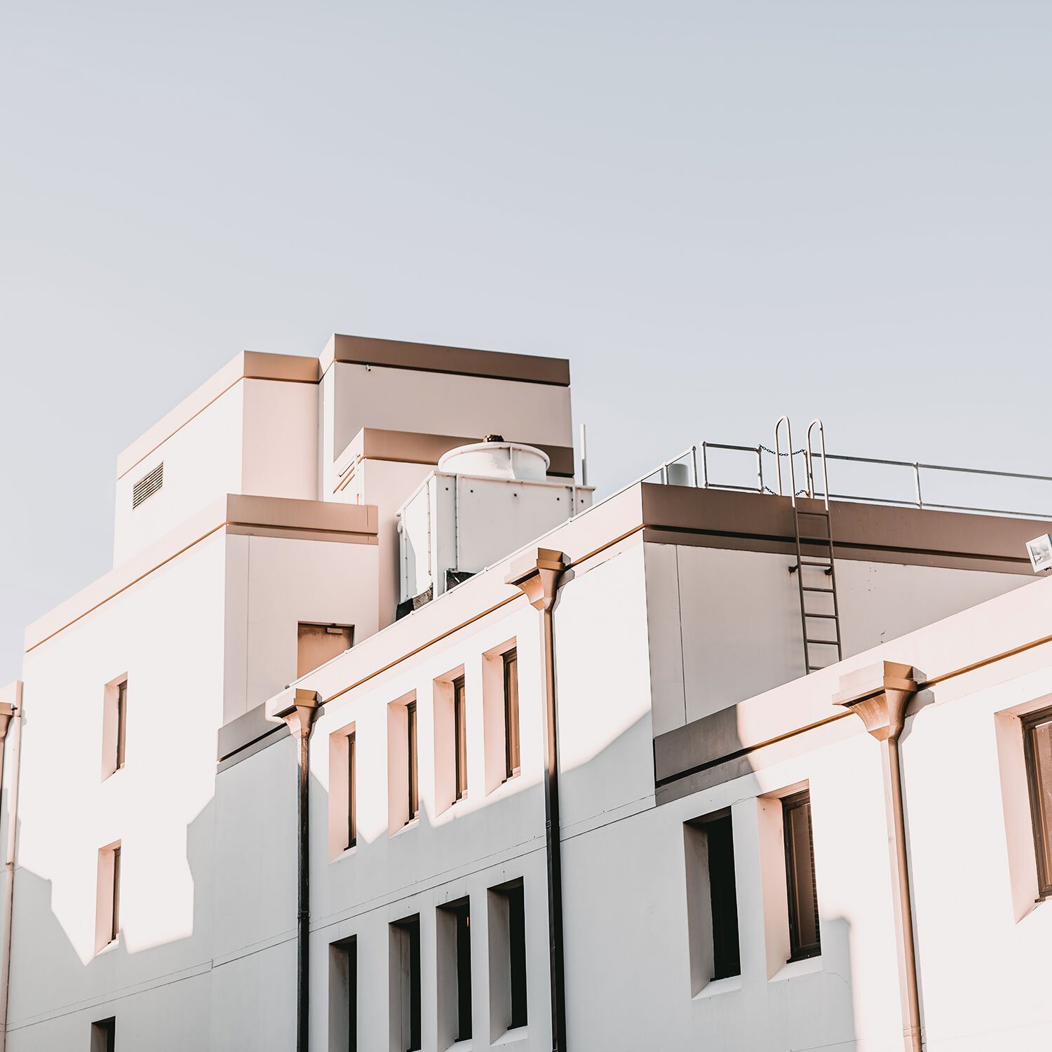Close-up of a multi-story white building with small windows, rooftop equipment, a ladder, and shadows cast on the wall, against a clear sky.