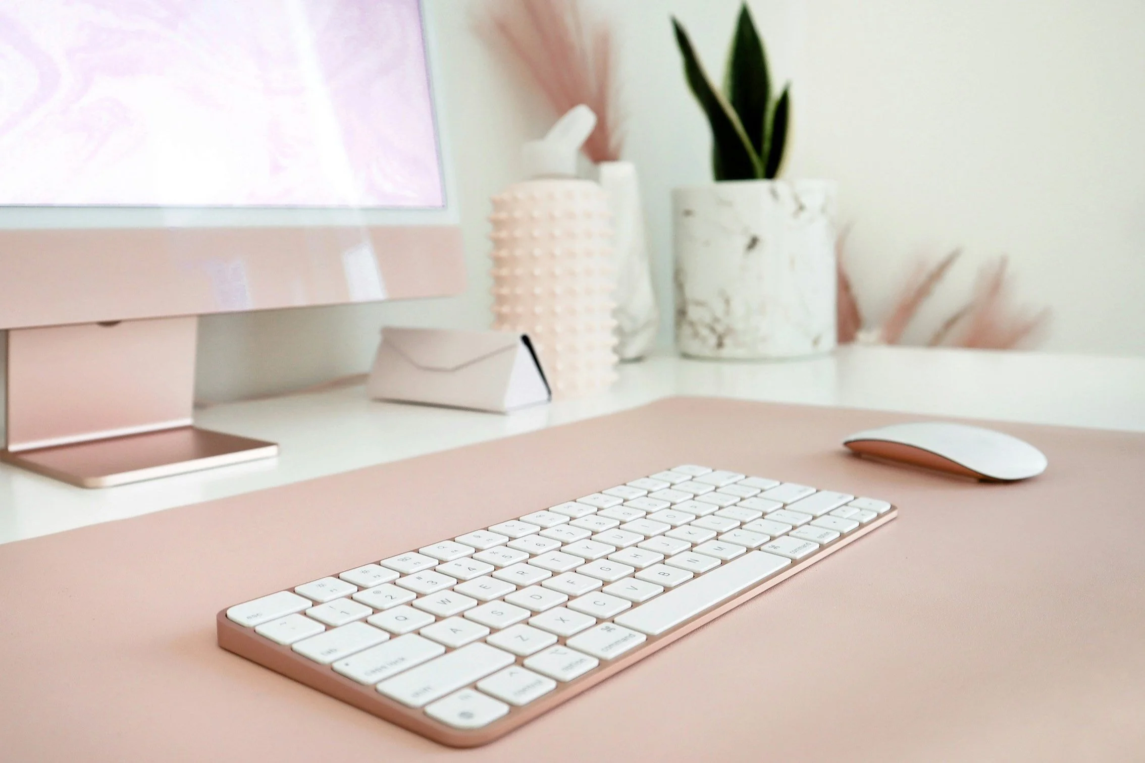 A pastel pink desk with a white keyboard and a white, curved computer mouse and a marble-patterned pot with a green plant. The background has soft pink and cream hues.