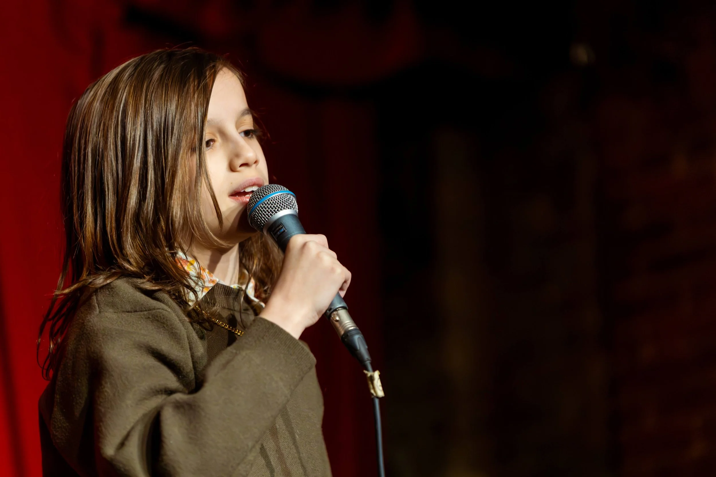 A young boy is holding a microphone and singing on stage. There are red curtains in the background. 