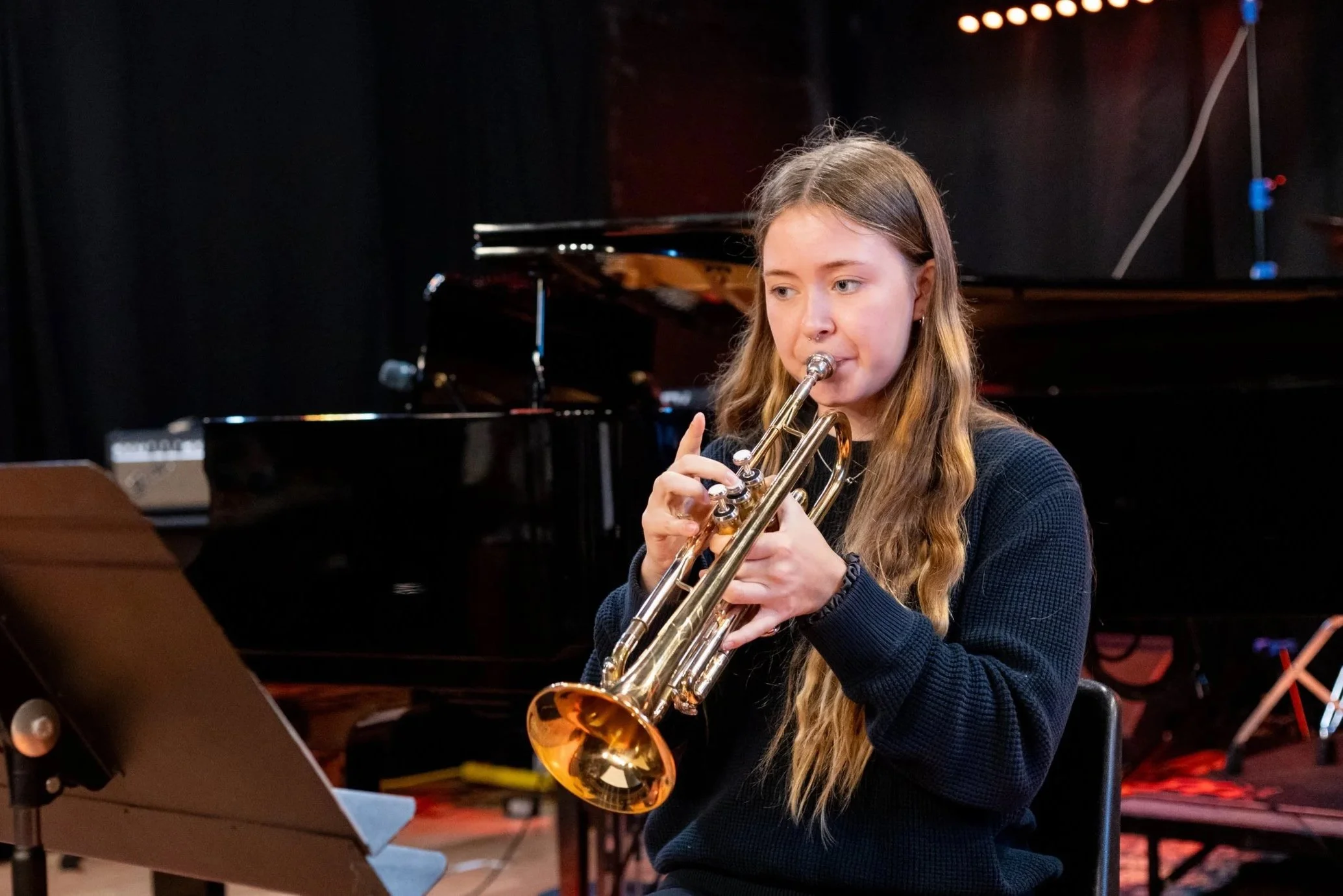 A teenage girl is playing trumpet on a stage. A grand piano is in the background.