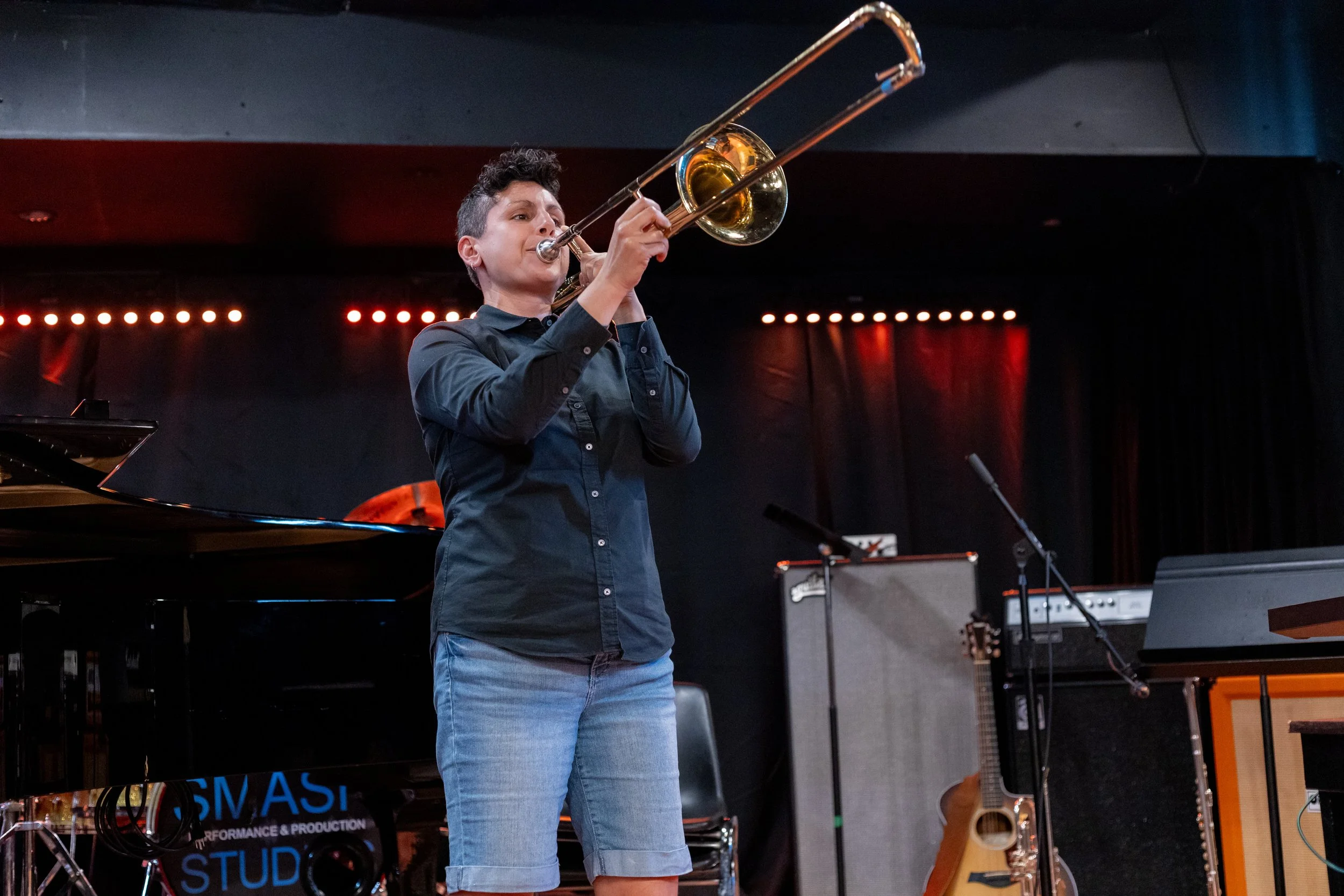 Female trombone player performing on stage at a concert. A guitar and a trumpet are in the background.