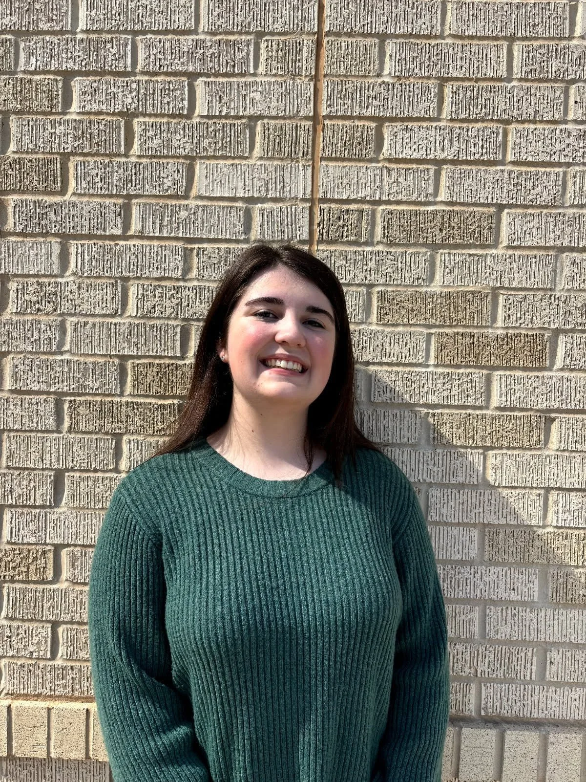 A young woman with dark brown hair smiling, wearing a green ribbed sweater, standing in front of a beige brick wall with a vertical metal pipe.