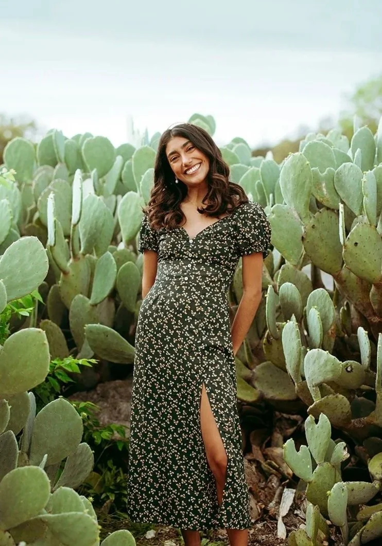A young woman with dark wavy hair wearing a black dress with white floral patterns standing among large green cactus plants, smiling at the camera.