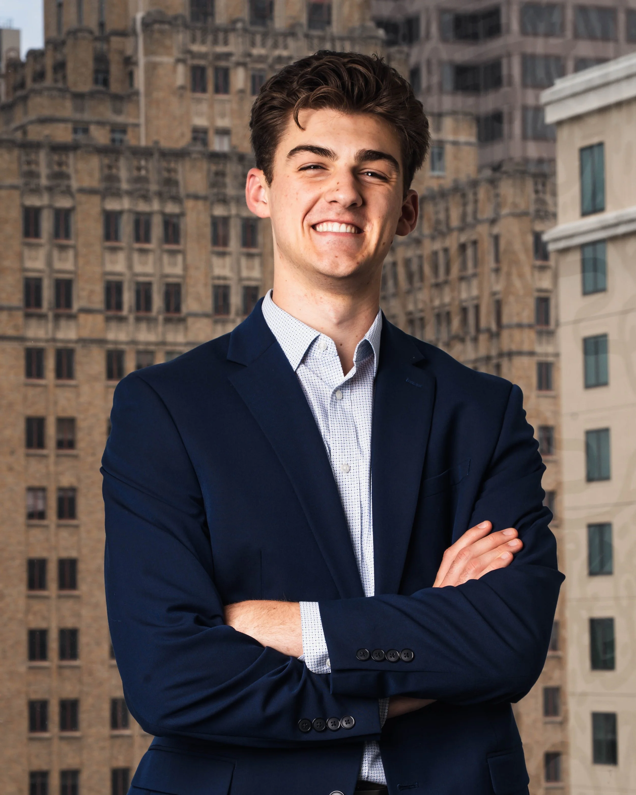 A young man with brown hair, smiling, wearing a navy blue suit and a white dress shirt, standing with arms crossed in front of a cityscape with tall buildings.