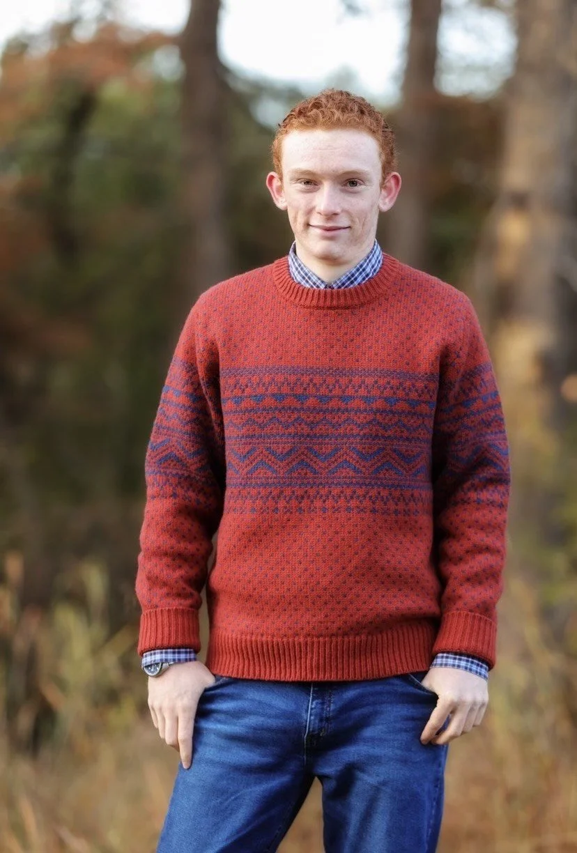 A young man with curly red hair wearing a patterned red sweater over a blue checkered shirt, standing outdoors with autumn foliage in the background.