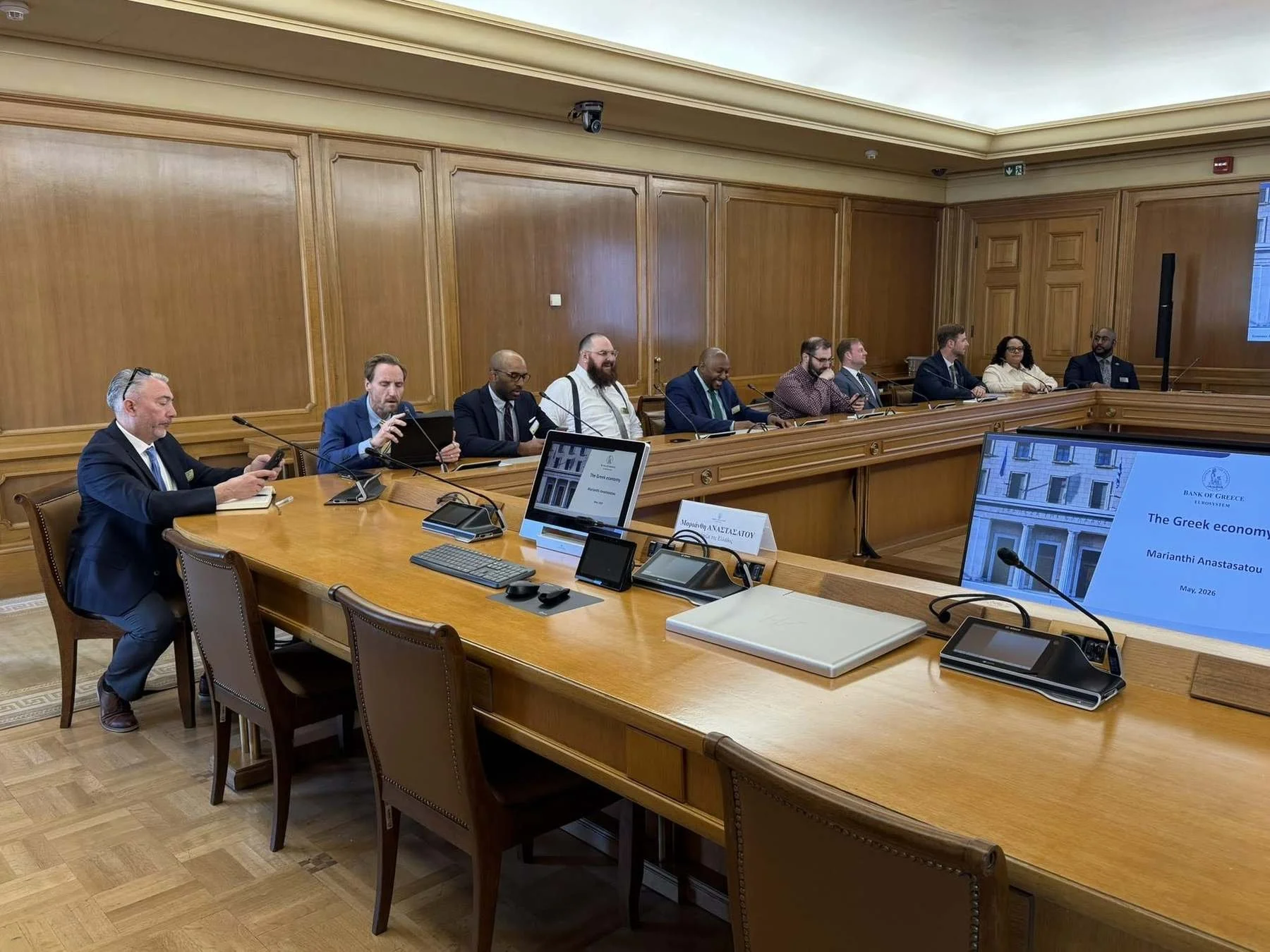 A briefing on the Greek economy at the Bank of Greece in Athens, delegates seated around a wood-panelled boardroom table facing a presentation screen.