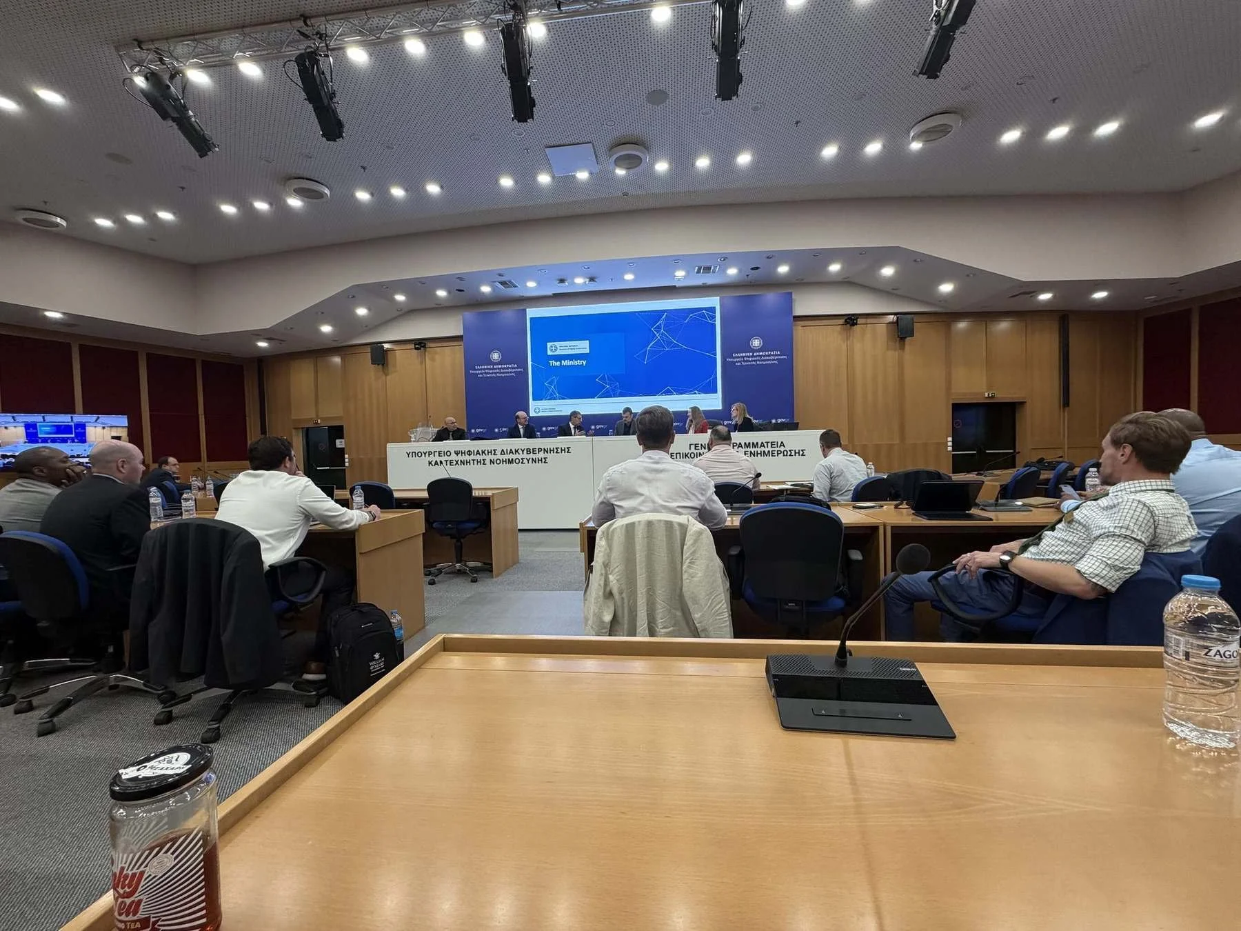 A briefing room at Greece's Ministry of Digital Governance and Artificial Intelligence in Athens, with the visiting delegation seated and a presentation slide reading The Ministry on the screen.