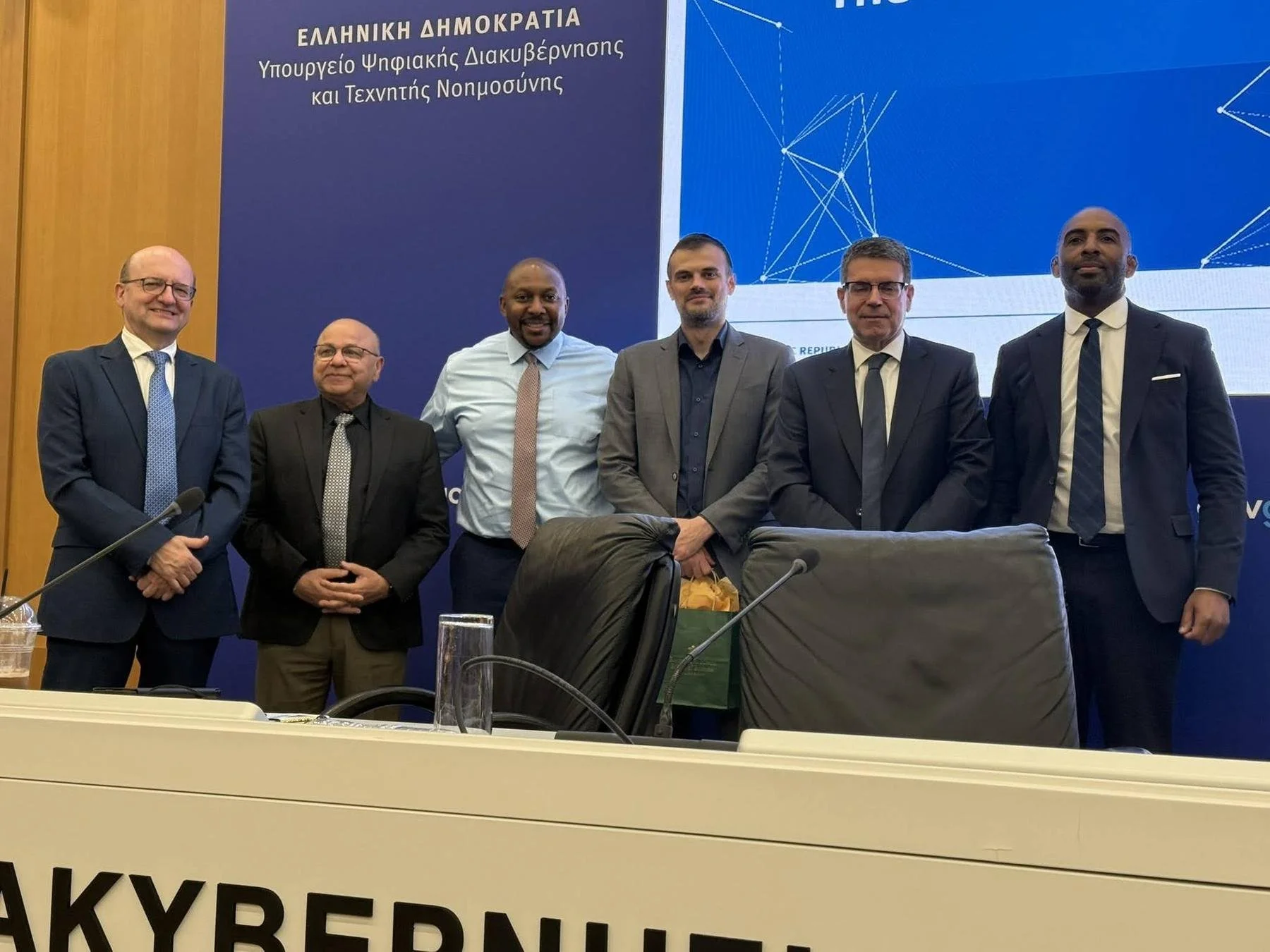 Six people standing together for a group photograph in front of the Hellenic Republic Ministry of Digital Governance and Artificial Intelligence banner in Athens.