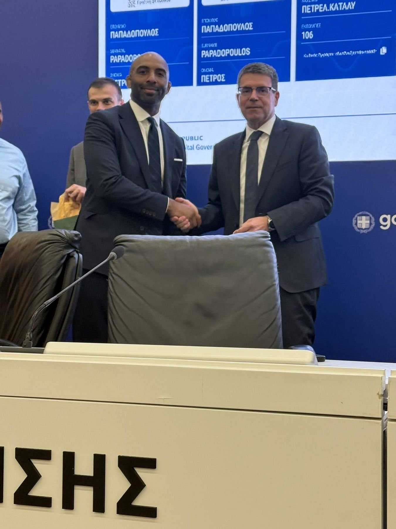 Two officials shake hands at a podium inside Greece's Ministry of Digital Governance and Artificial Intelligence, with a gov.gr record displayed on the screen behind them.