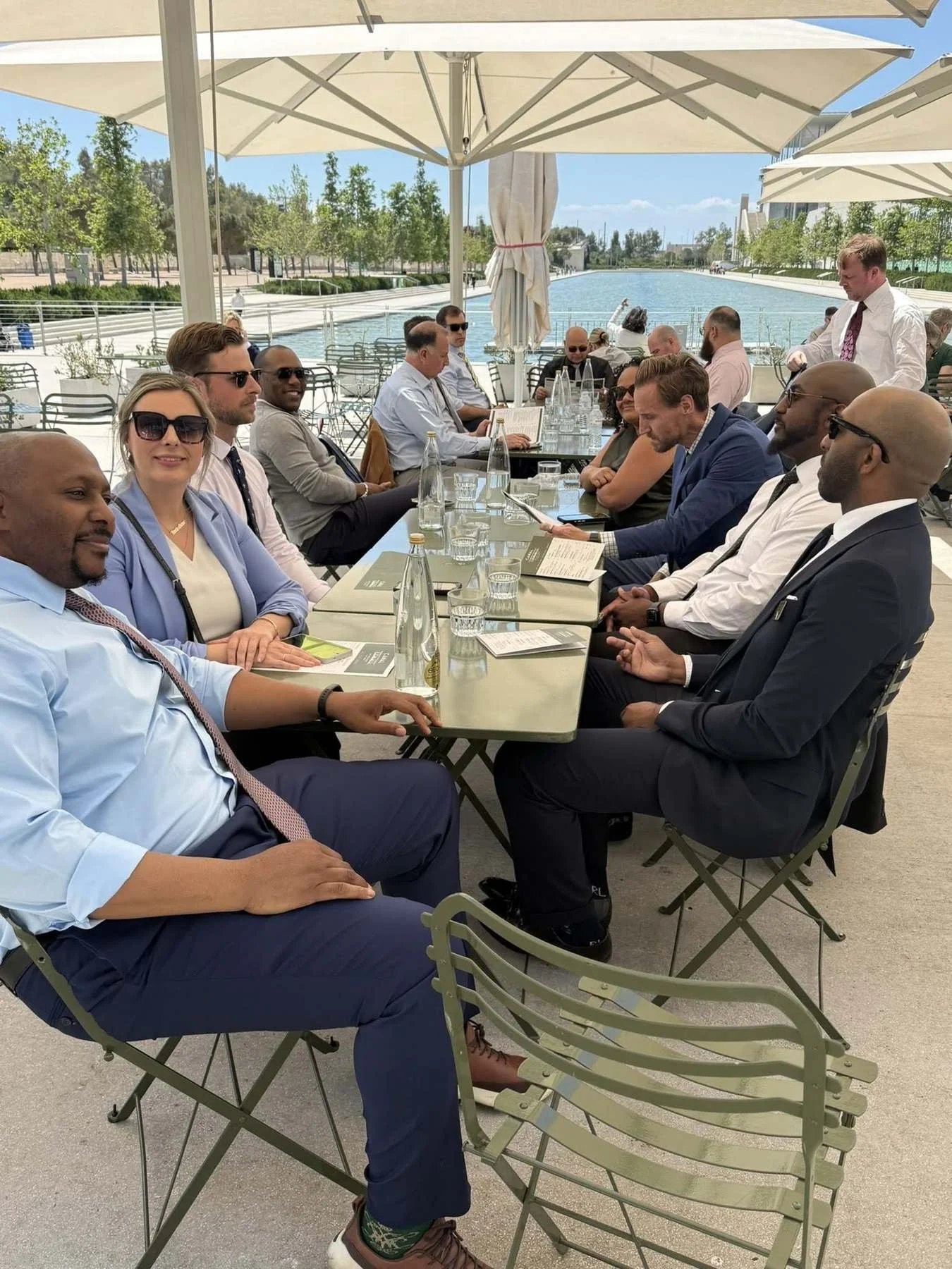 The delegation at a long outdoor lunch table under white umbrellas beside a canal in Athens on a clear day.