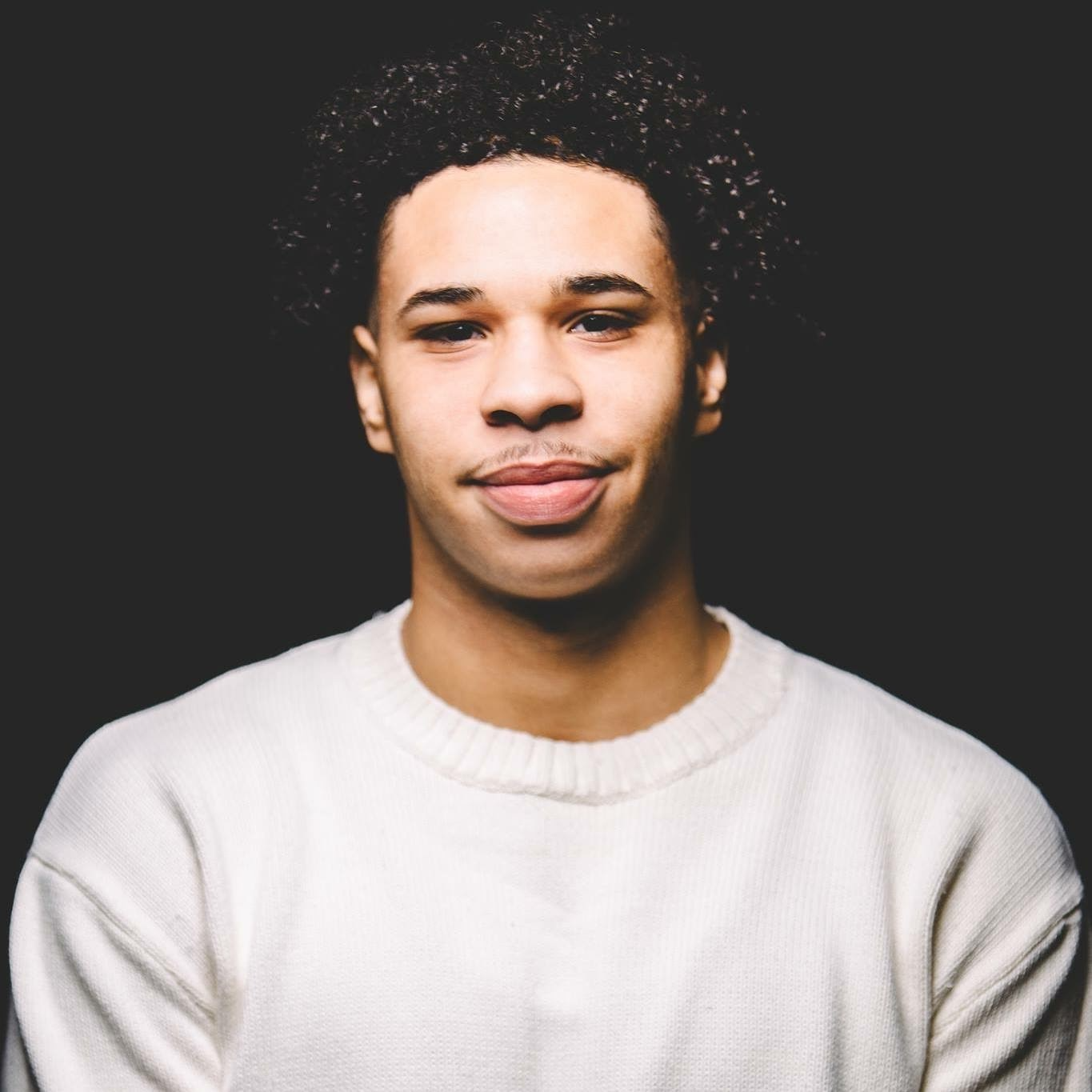 Portrait of a young man with curly hair wearing a white sweater against a black background.