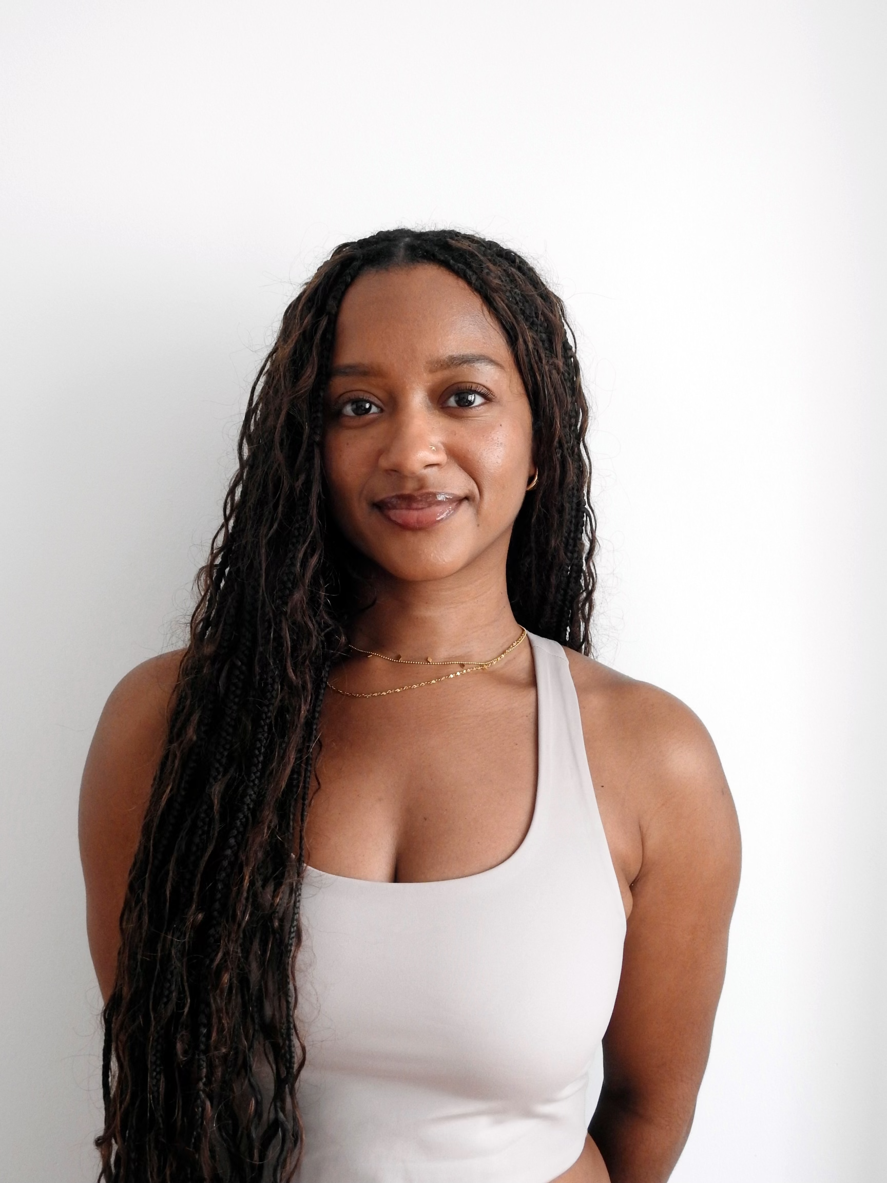 Portrait of a young woman with long twists, wearing a light gray tank top and gold jewelry, standing against a plain white wall.