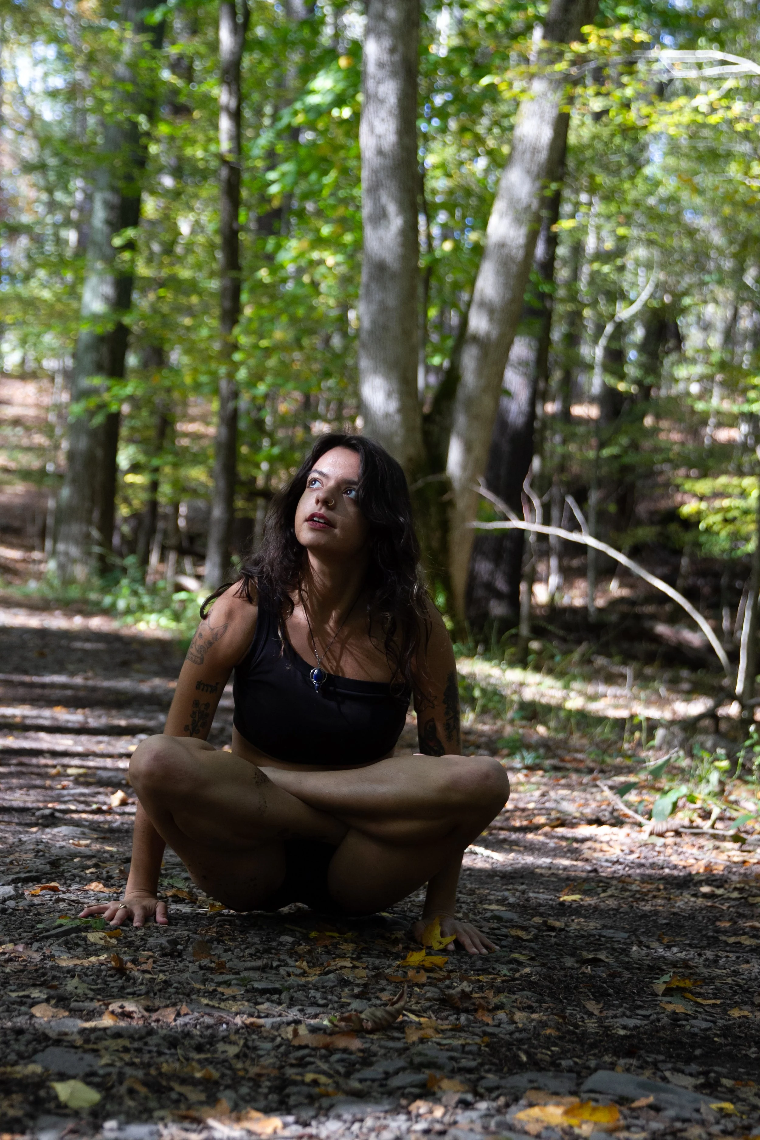 A woman with dark wavy hair and tattoos on her arms is sitting cross-legged on the forest ground, wearing a black crop top with leaves and trees in the background.