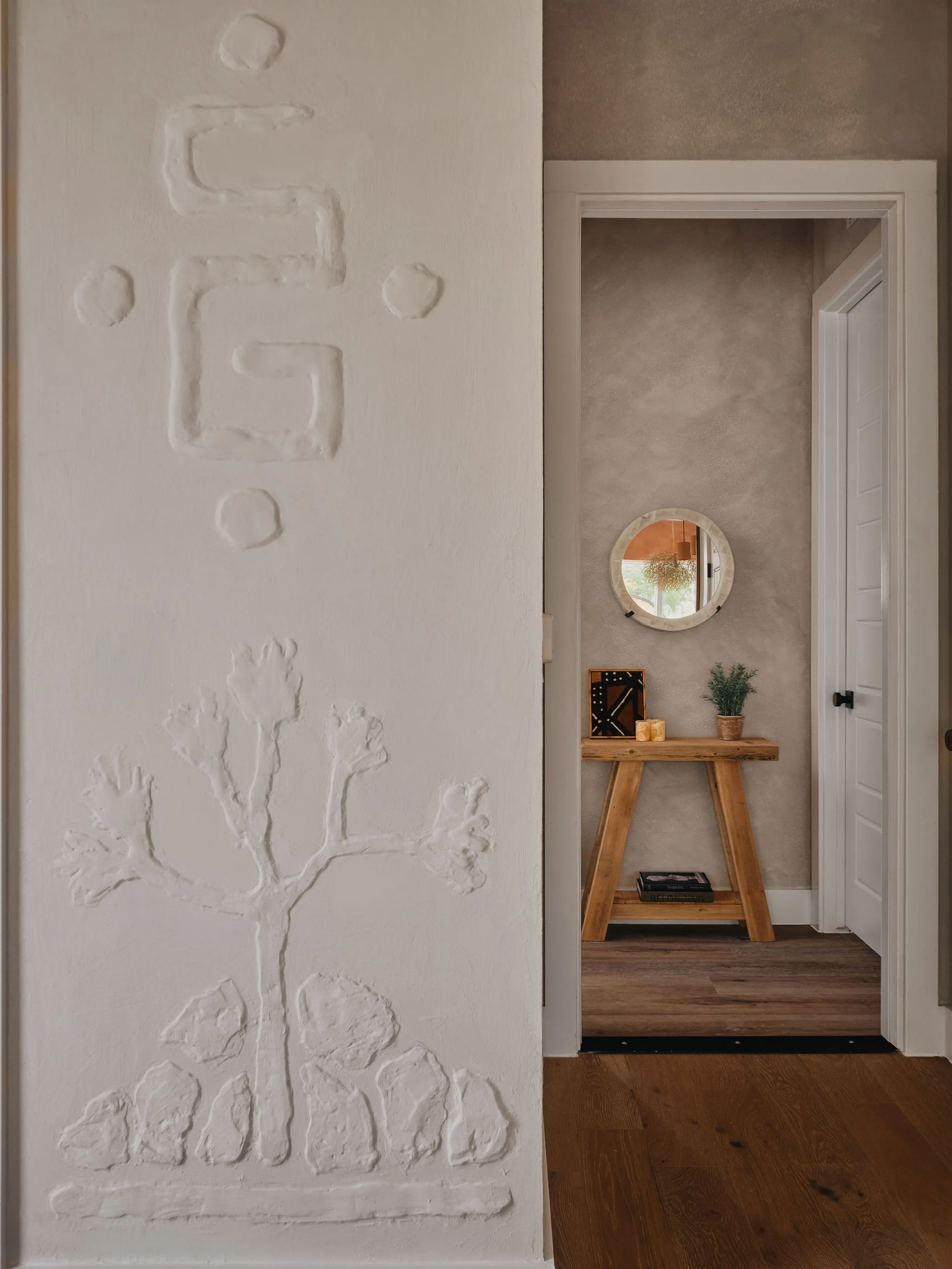 A wall with textured, embossed artwork and a view into a room with a wooden console table, circular mirror, plants, candles, and a window.