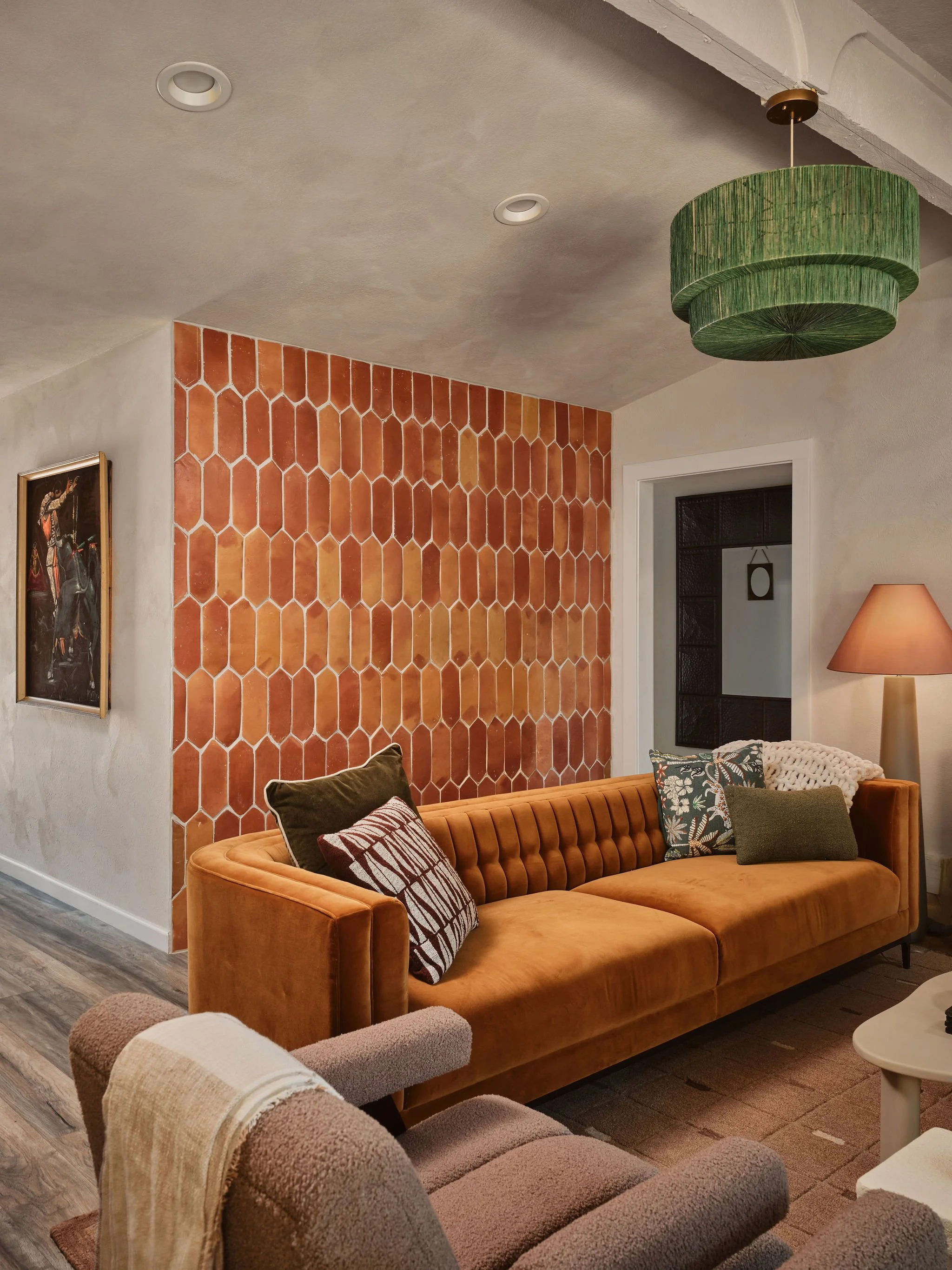 A cozy living room with an orange tufted velvet sofa, decorative pillows, a green hanging lamp, a terracotta hexagonal tile accent wall, a framed painting, and a beige upholstered armchair with a throw blanket. The room has a doorway with a white fra