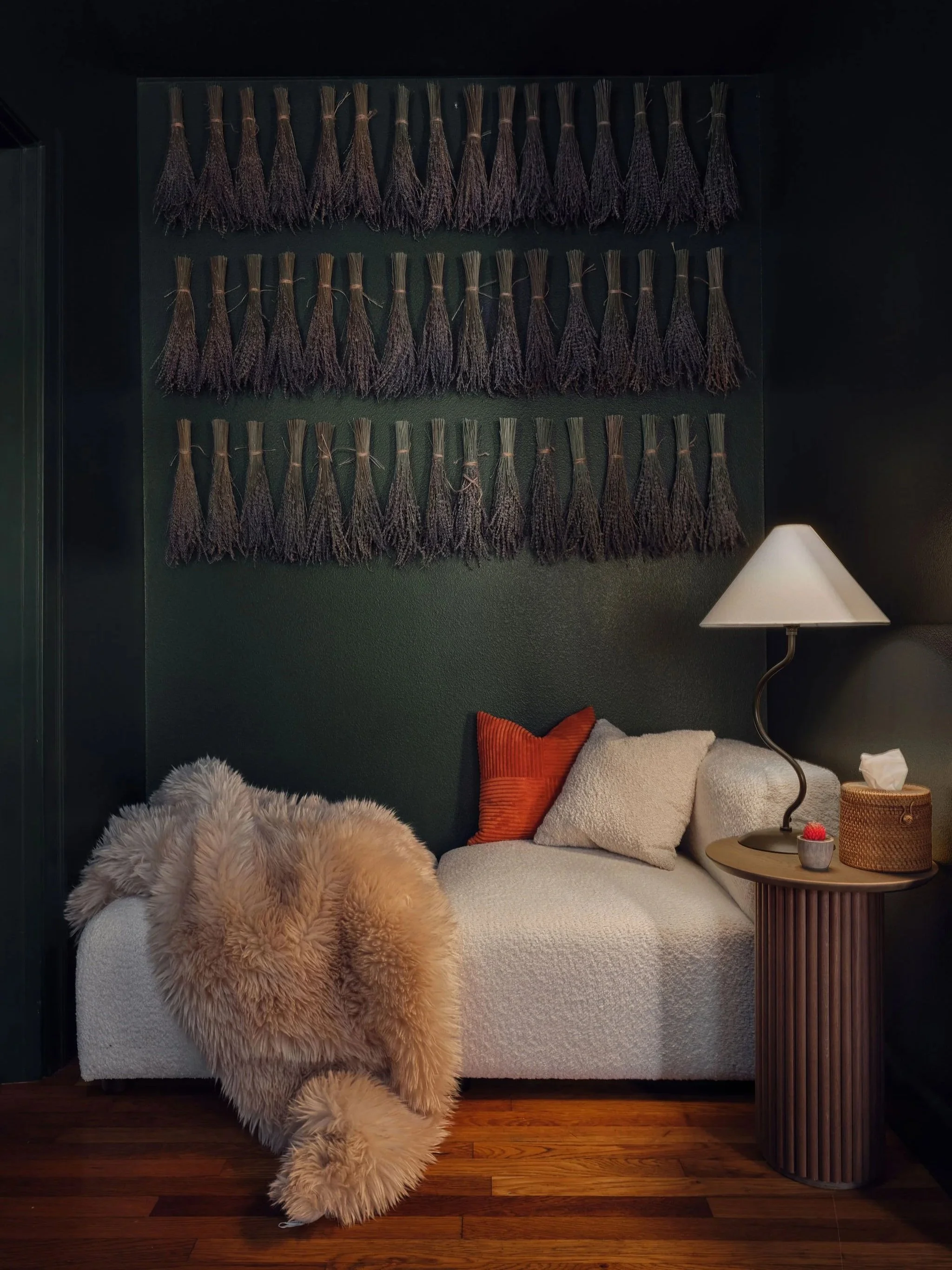 A living room corner with a white sofa, fluffy beige throw blanket, orange pillow, beige cushion, a wooden side table with a lamp, tissue box, and small bowl, dark wall with three rows of lavender bundles hanging, wooden floor.