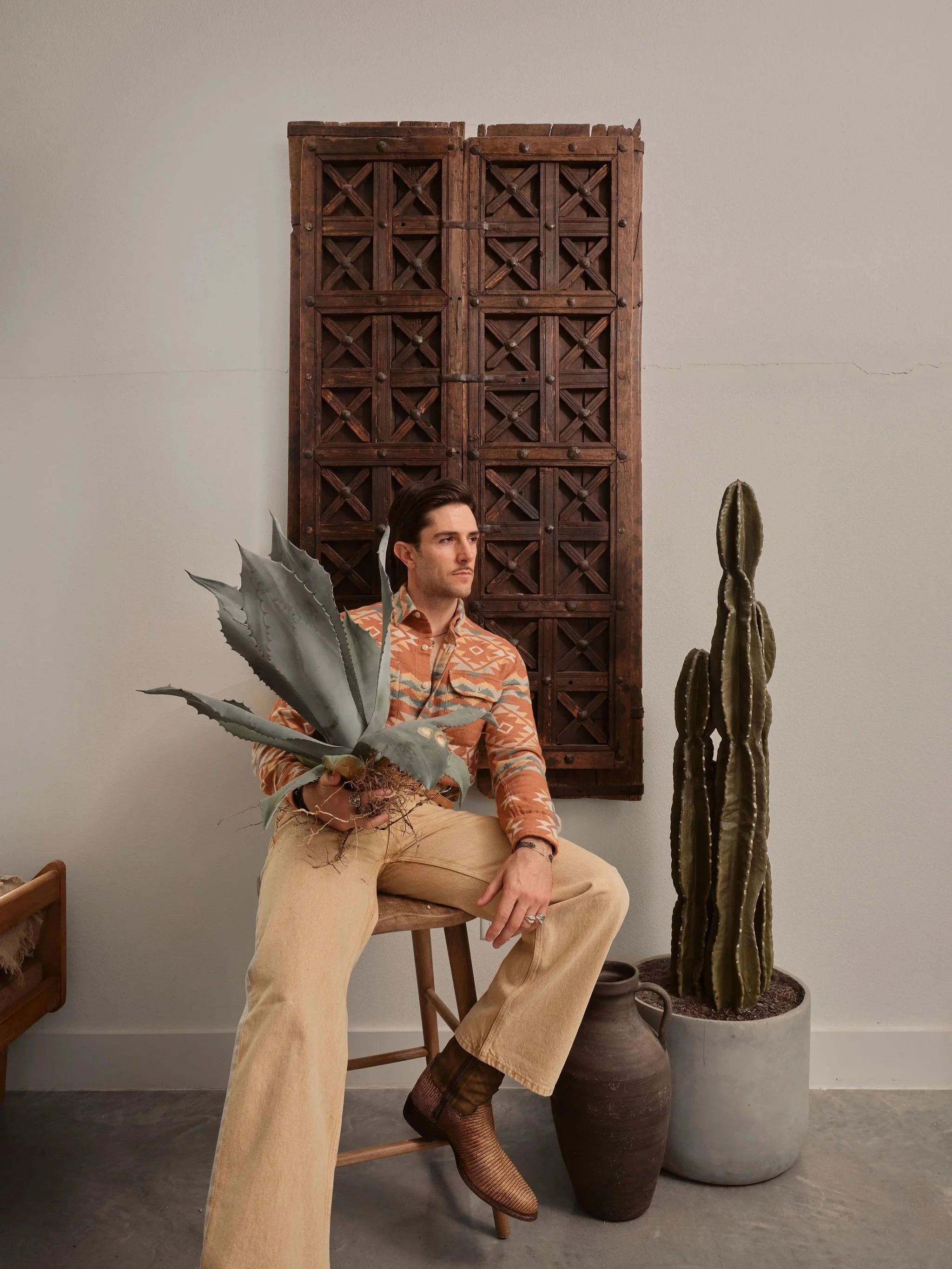 A man sitting on a stool holding a large potted agave plant, with a cactus in a large pot and wooden wall hanging in the background.
