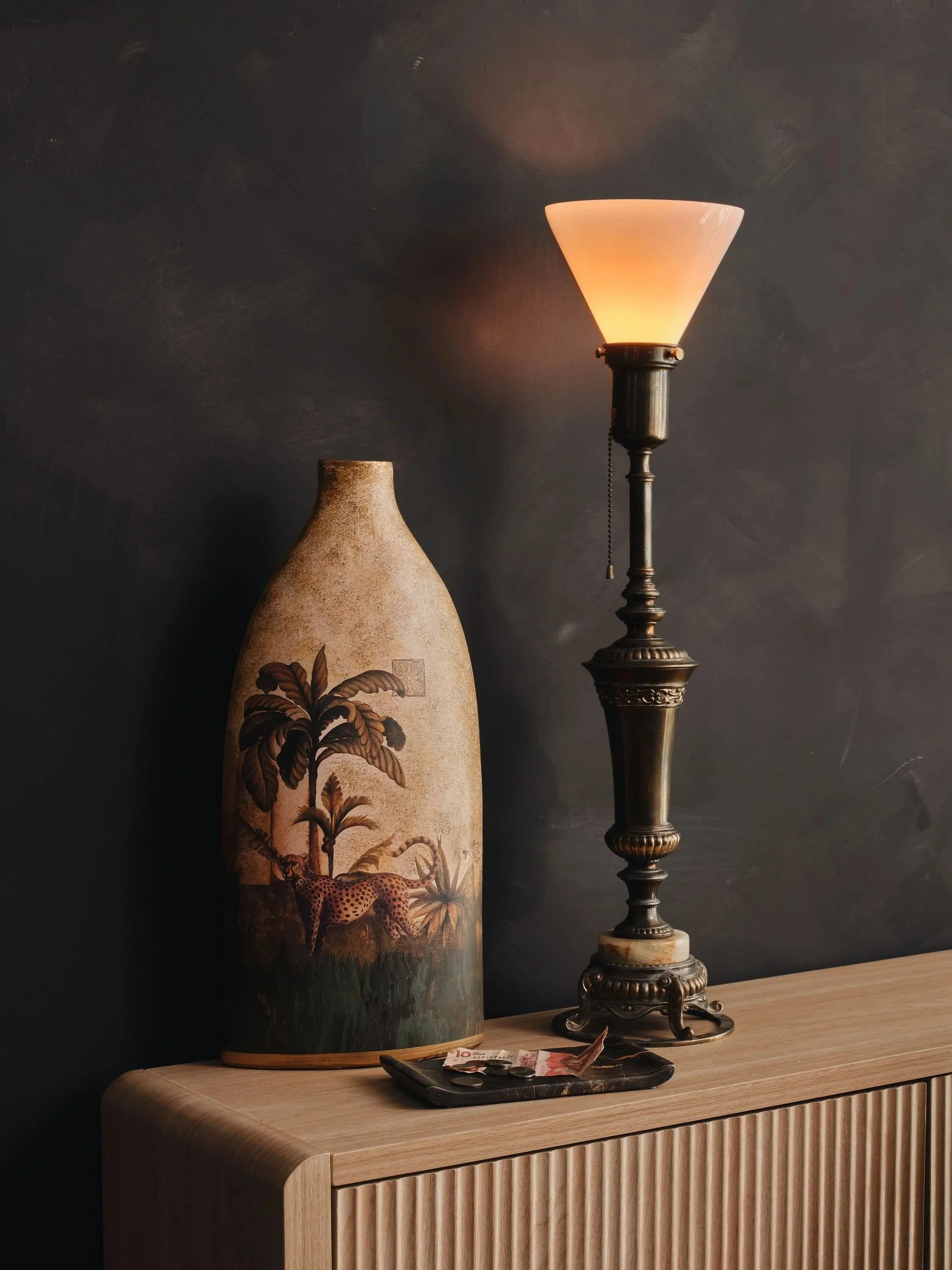 A decorative table lamp with a beige shade on a dark stand next to a tall ceramic vase with a painted scene of trees, a leopard, and a butterfly. There is a tray with a few coins and paper currency on the light wood surface in front of the vase and l