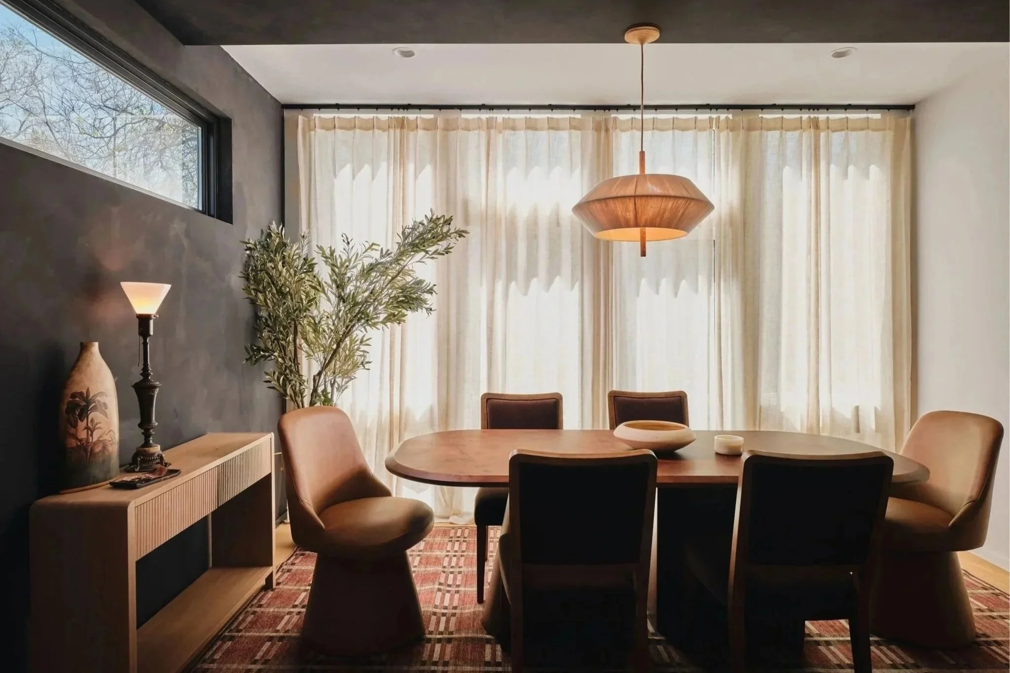 Interior of a dining room with a round wooden table surrounded by beige upholstered chairs, large cream curtains on windows, a modern pendant light hanging over the table, a sideboard with decorative vases and a lamp, and a patterned rug on the floor