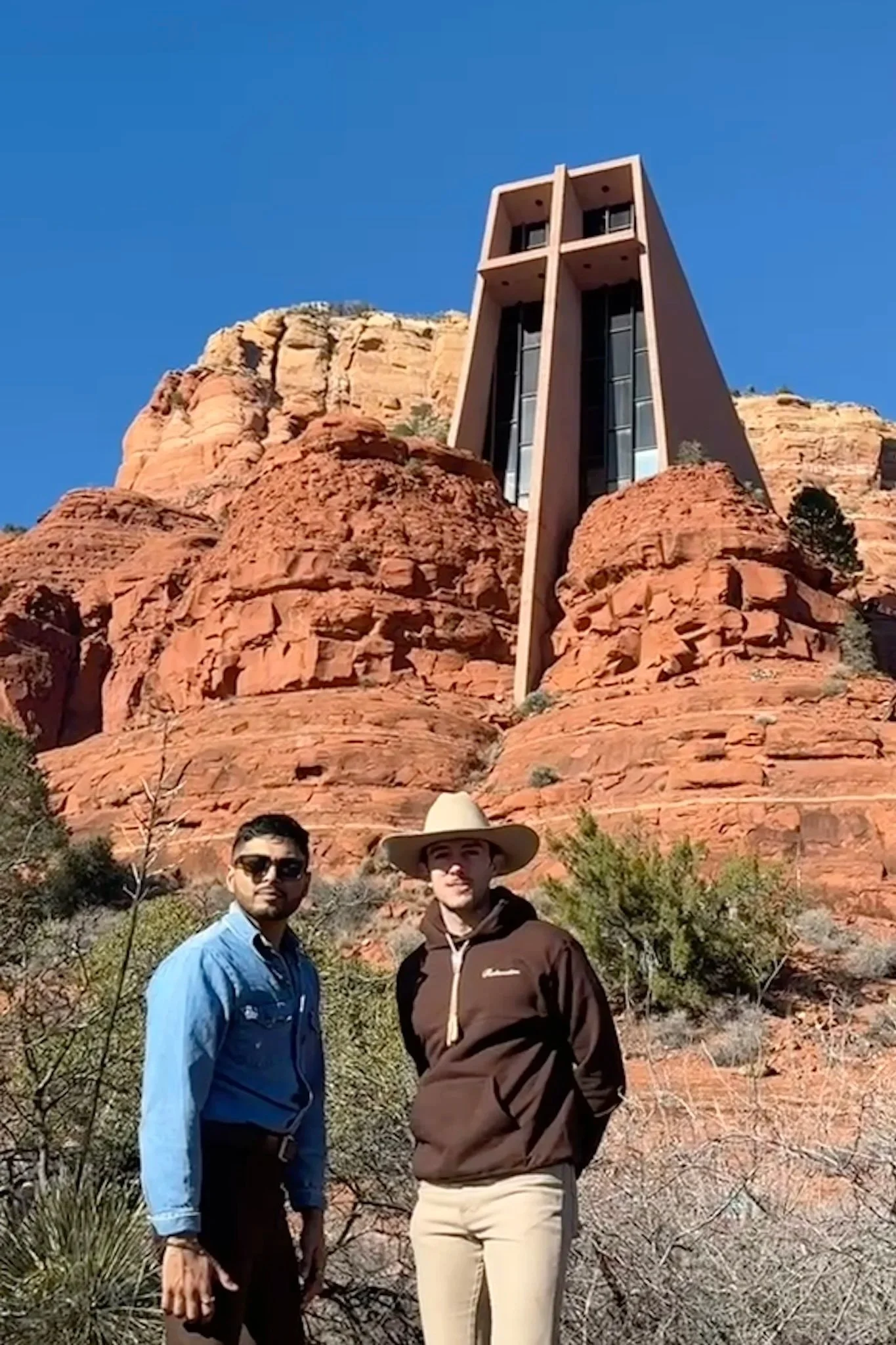 Two men standing in front of red rock formations and a modern building on top of the cliffs under a clear blue sky.