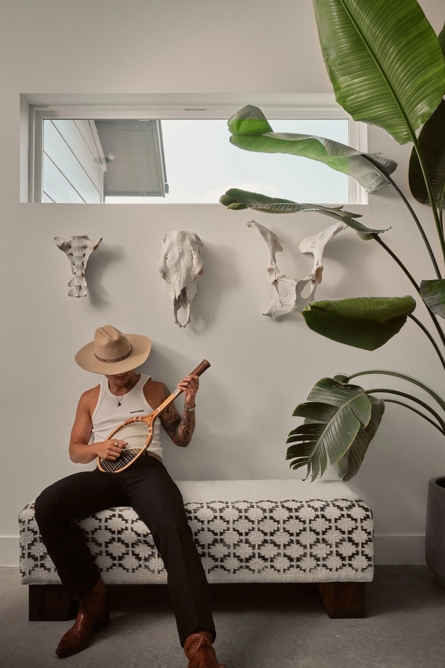 A person wearing a straw hat, white tank top, and black pants, sitting on a patterned bench, holding a tennis racket, and looking down, with a large green plant and white animal skull decorations on the wall behind them.