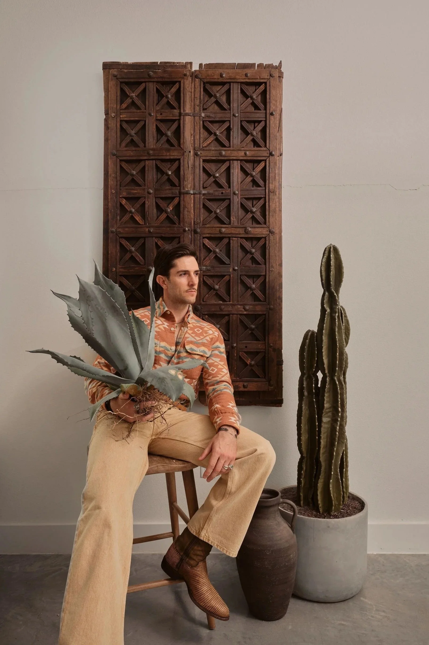 A man sitting on a stool holding a large potted agave plant, with a cactus in a large pot and wooden wall hanging in the background.