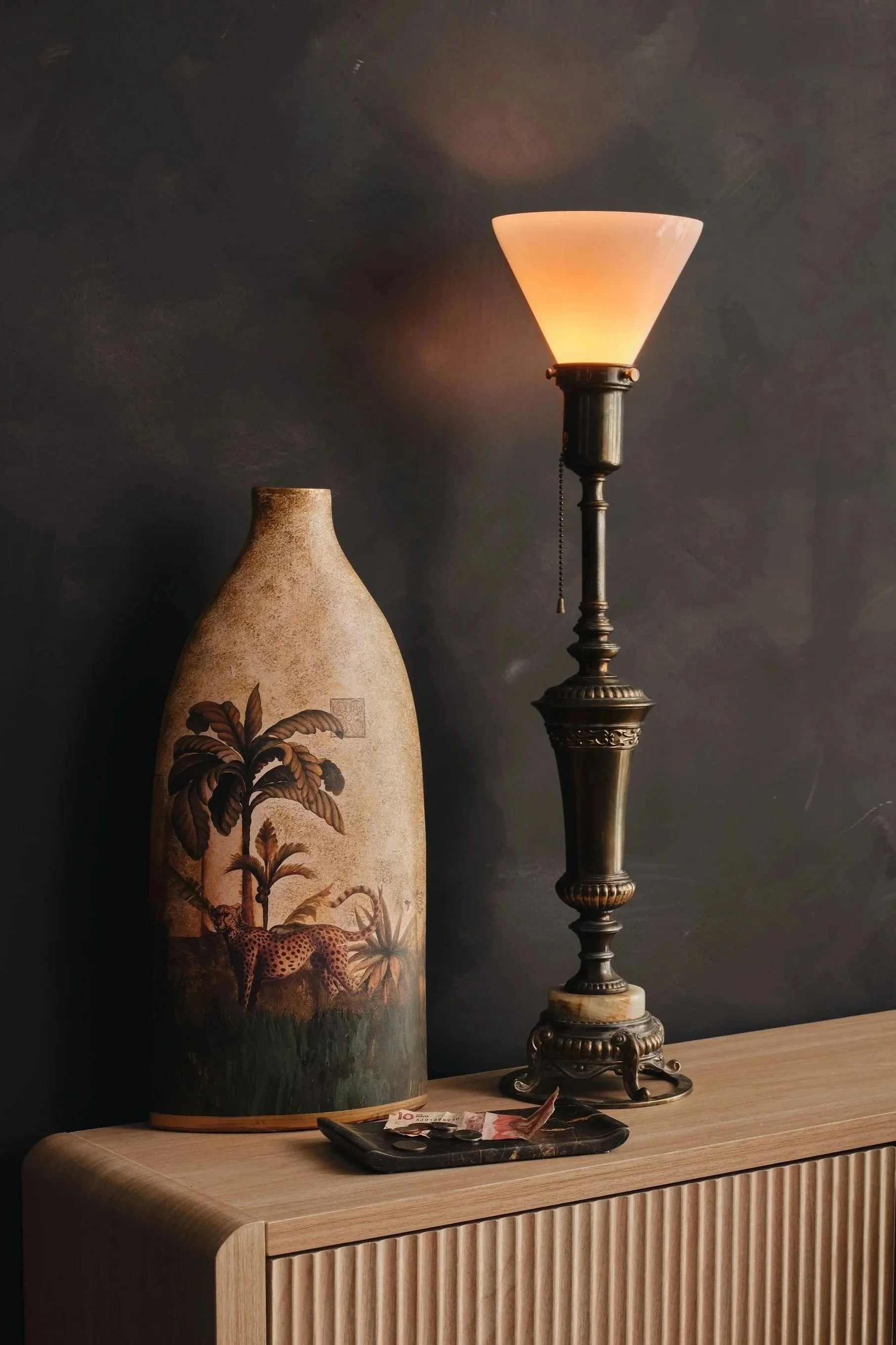 A decorative table lamp with a beige shade on a dark stand next to a tall ceramic vase with a painted scene of trees, a leopard, and a butterfly. There is a tray with a few coins and paper currency on the light wood surface in front of the vase and l