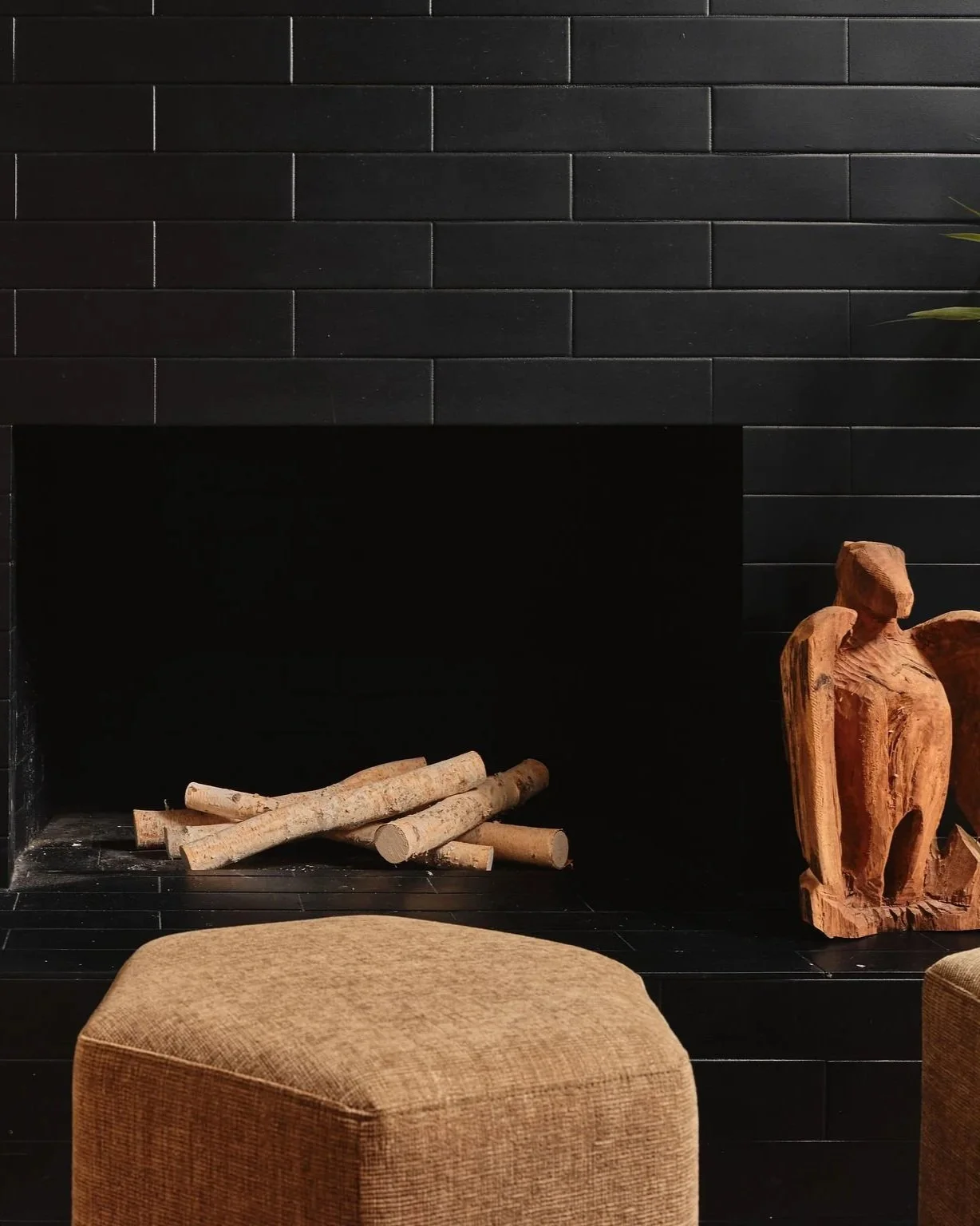 Black brick fireplace with a pile of firewood on the hearth and a carved wooden sculpture beside it, with a beige cushioned stool in the foreground.