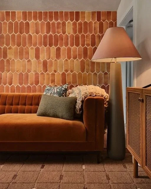 Living room scene featuring an orange velvet sofa with decorative pillows, a tall beige floor lamp with a cone-shaped shade, and a brown wood cabinet against a honeycomb-patterned terracotta wall.