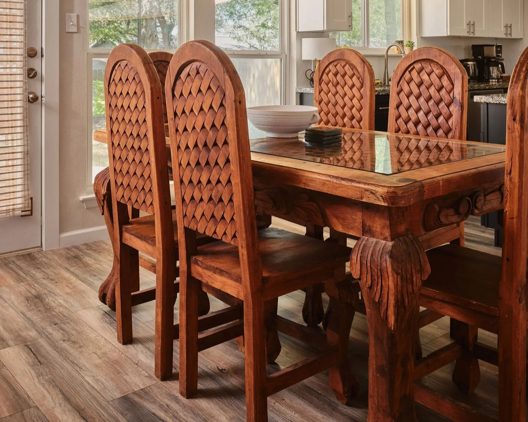 A wooden dining table with six matching high-backed chairs in a kitchen, with a large bowl and a set of coasters on the table, and sunlight coming through large windows in the background.