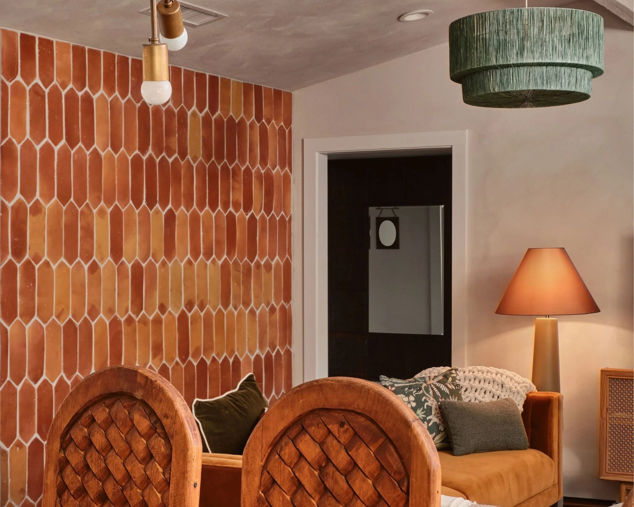 Interior room with orange hexagonal tile wall, three wooden chairs, a green pendant lamp, a table lamp with a beige shade, and a patterned cushion.