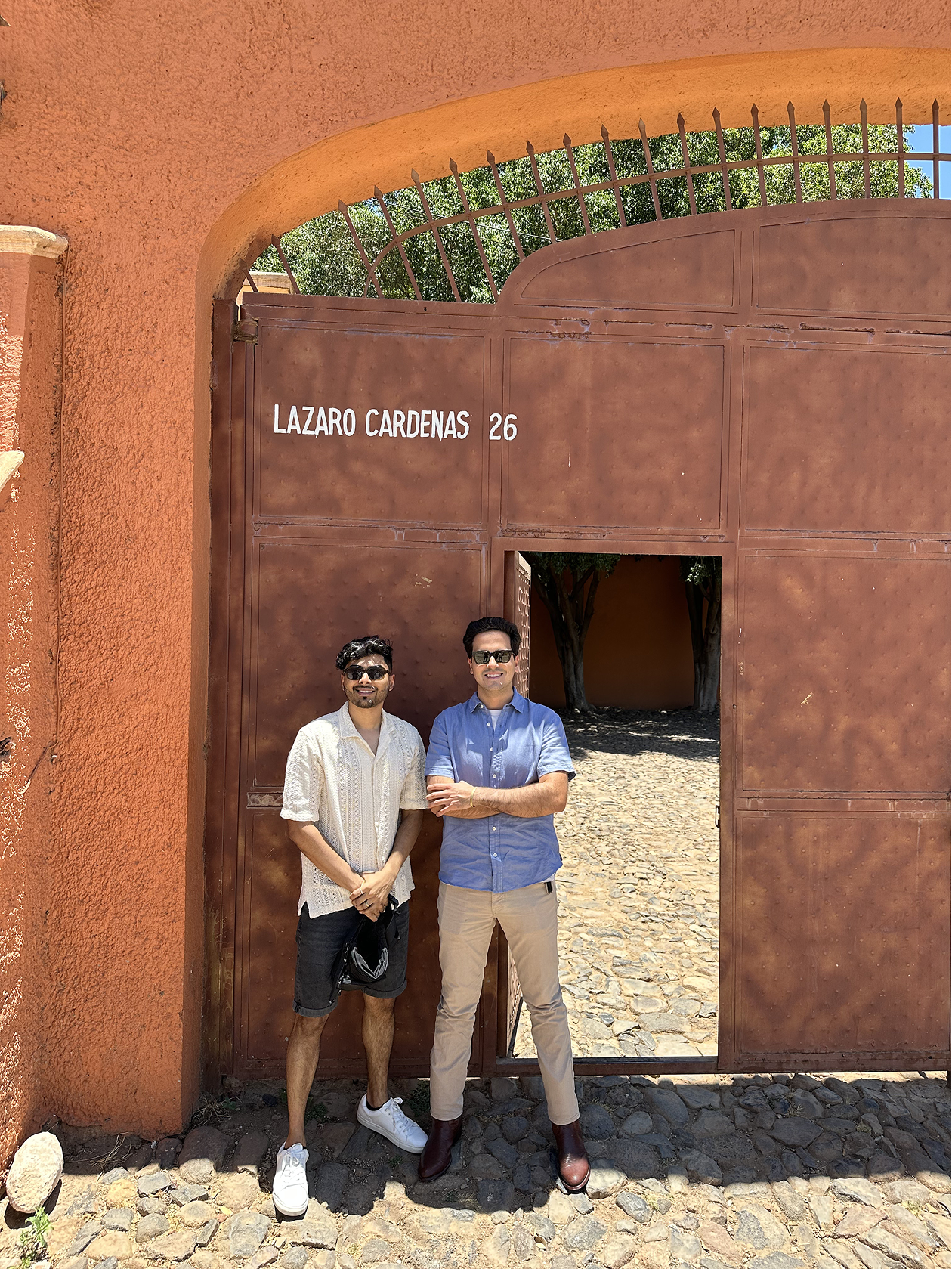 Two men standing in front of a large gate with the name 'Lazaro Cardenas 26'. They are outdoors, under sunlight, with trees visible through the gate opening.
