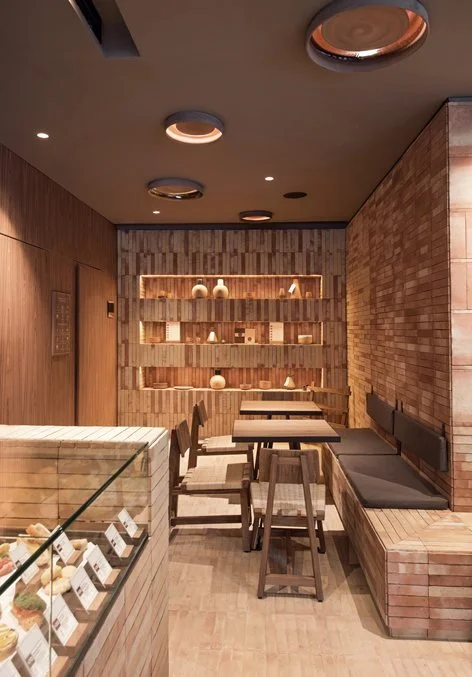 Interior of a modern café with wooden accents, tables, chairs, a brick wall, and display shelves with decorative items.