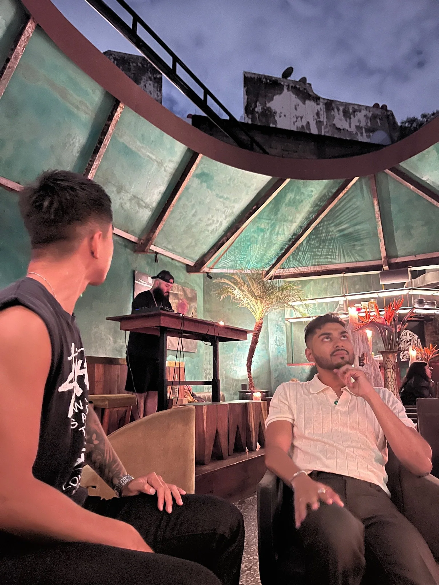 People sitting in a restaurant with tropical decor, including a large palm tree painting, and a DJ playing music in the background.