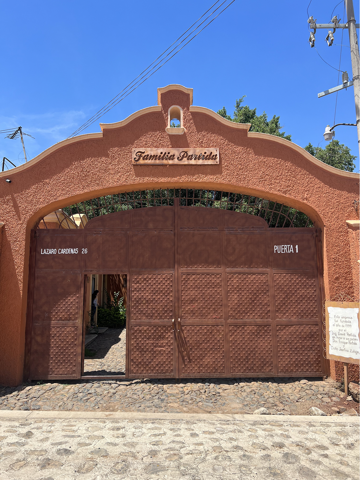 Photo of an entrance gate to a property called Familia Partida with a sign that reads Lazo Cardenas 26 and Puerta 1. The gate is made of metal and painted brown, with a textured pattern. The surrounding wall is pinkish-orange stucco with decorative edges. There is a small sign or plaque to the side providing additional information about the property, which appears to be a family estate or commemorative site. The background shows a bright blue sky and some trees.