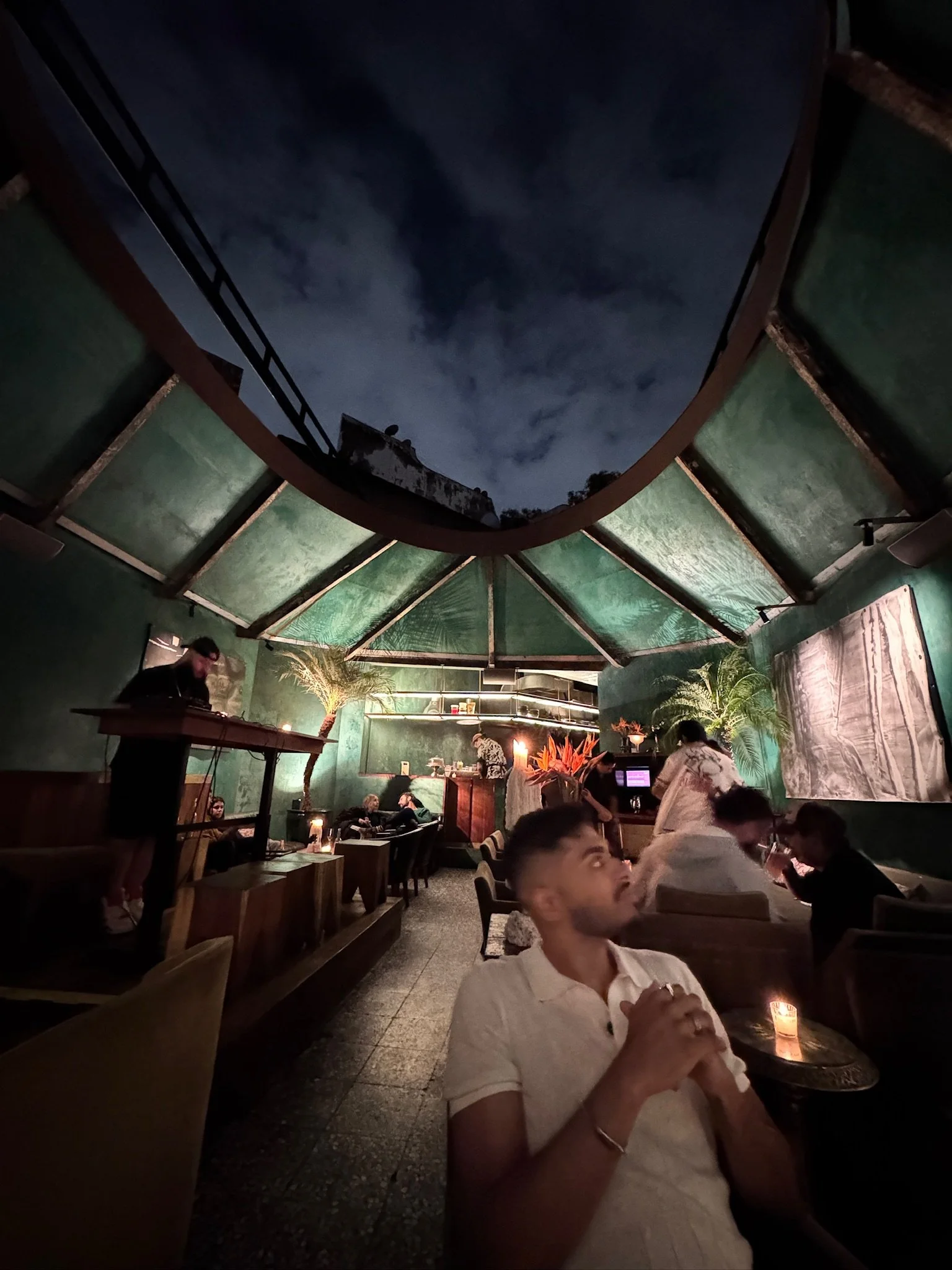 Interior of a dimly lit restaurant with a high ceiling and a mural on the wall. People are seated at tables, some are eating and talking. A man in the foreground is holding a drink, and a woman nearby is drinking. There are decorative plants and candles creating a cozy atmosphere.