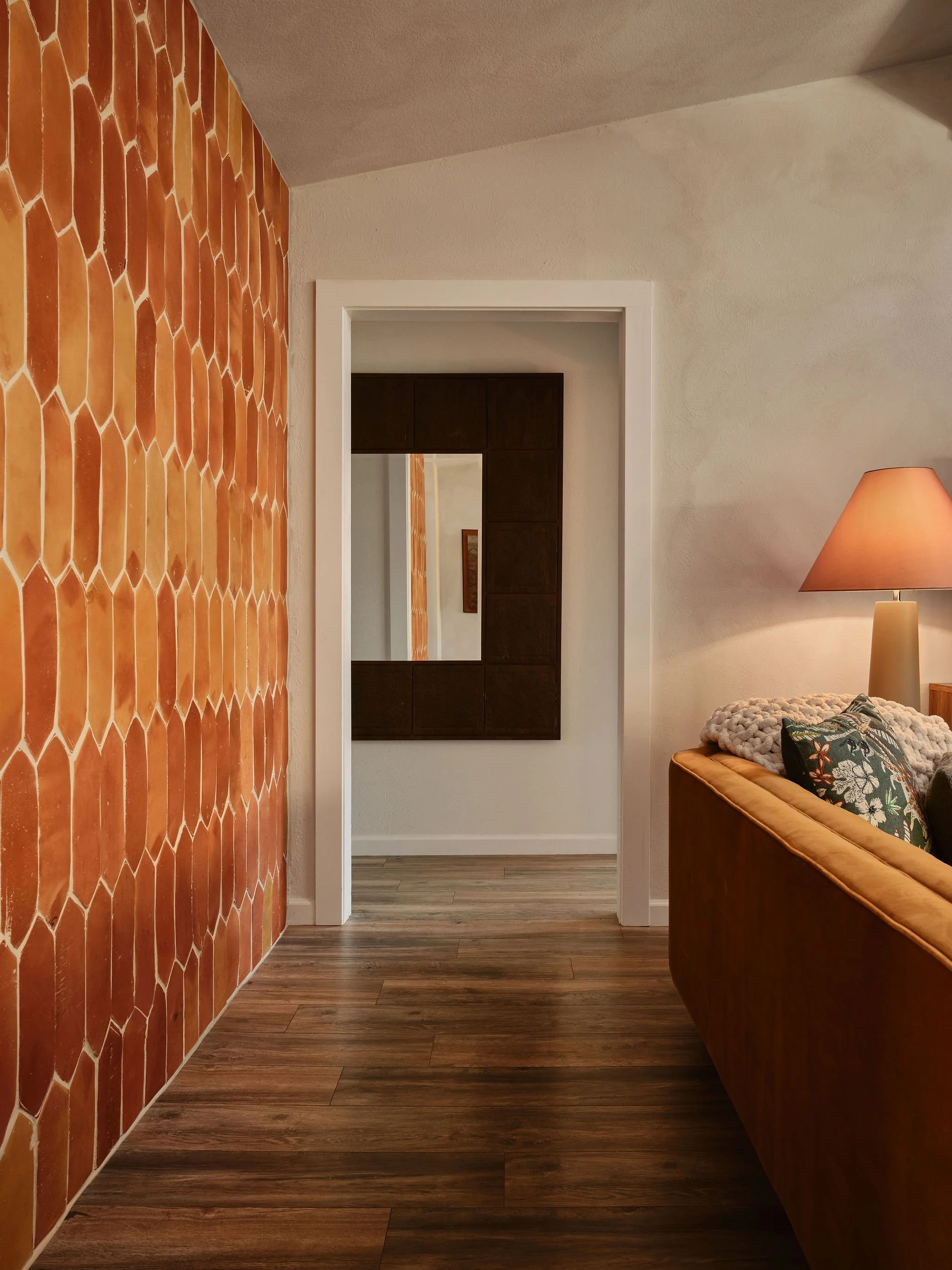 View of a room with wood flooring, a wall on the left with orange terracotta tiles, a beige textured wall in the background with a mirror and wooden cabinet, and a portion of a tan sofa with patterned pillows and a table lamp on the right.