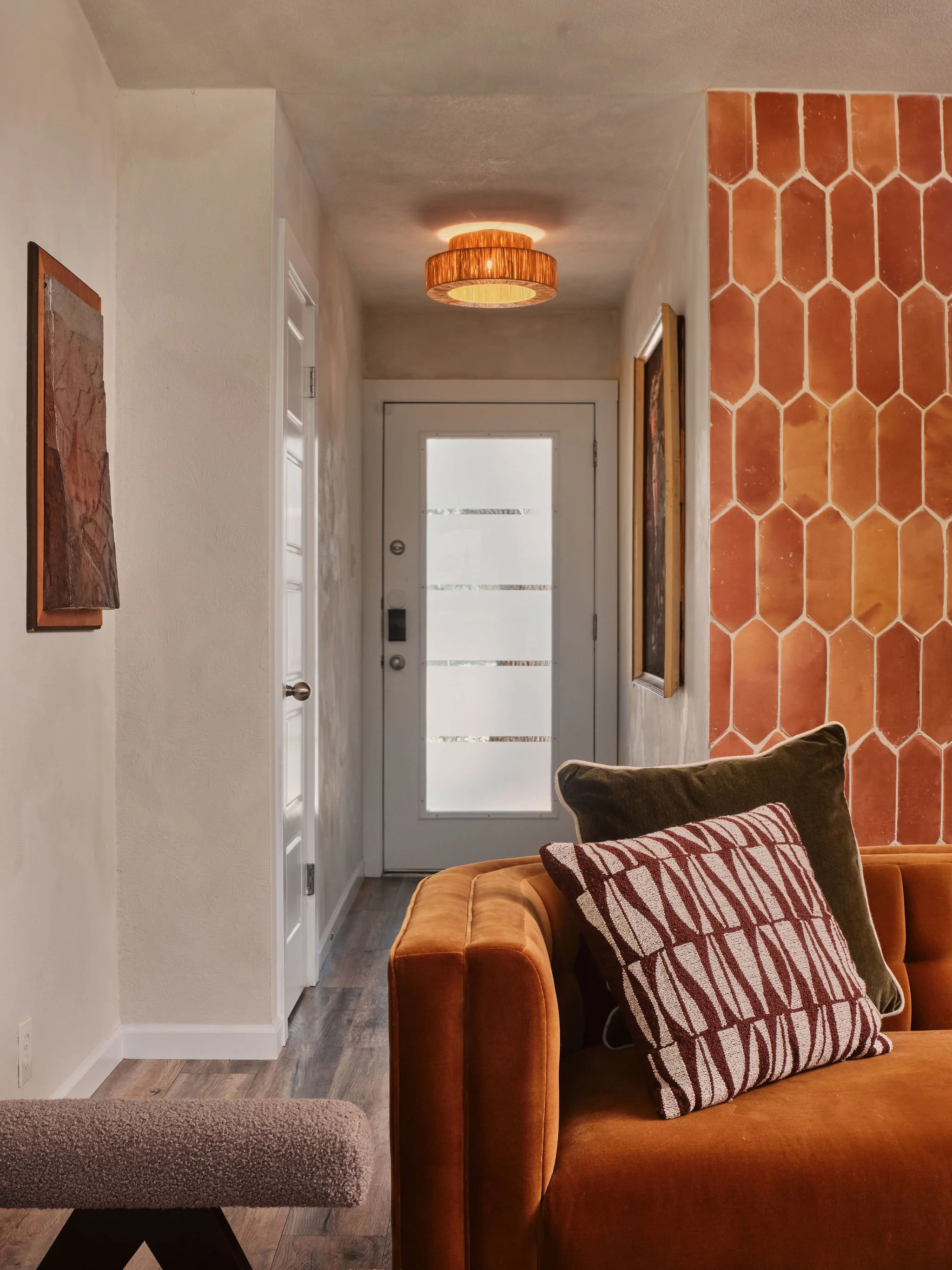 A cozy living room with a mustard yellow sofa, striped pillows, and a terracotta tile accent wall near the entry door. The entry door has horizontal glass panels, and there is a ceiling light fixture with a wooden shade. Part of a beige chair is visi