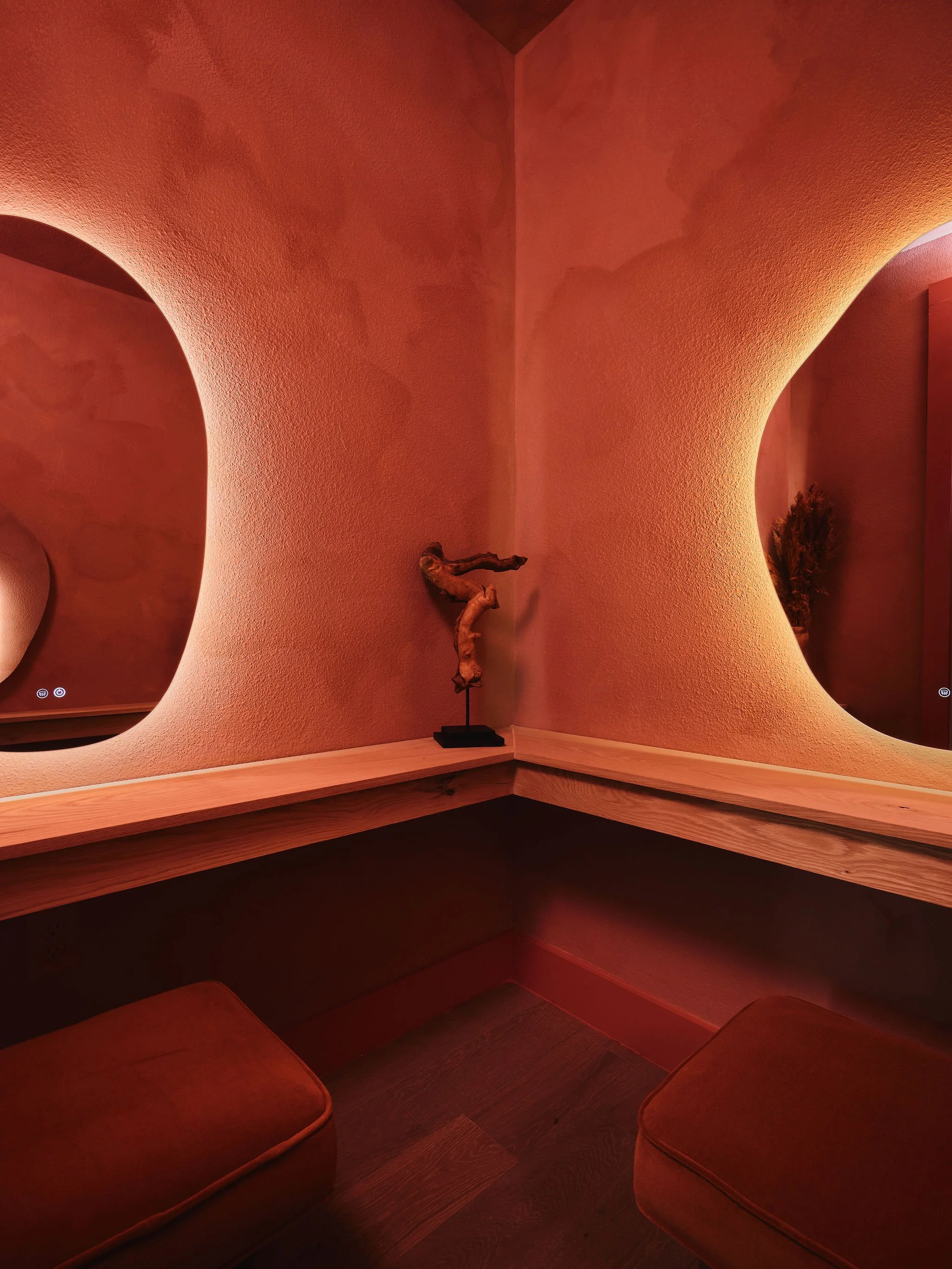 Cozy corner with red cushioned seating, a small wooden decorative branch, a curved mirror, and subtle warm lighting.
