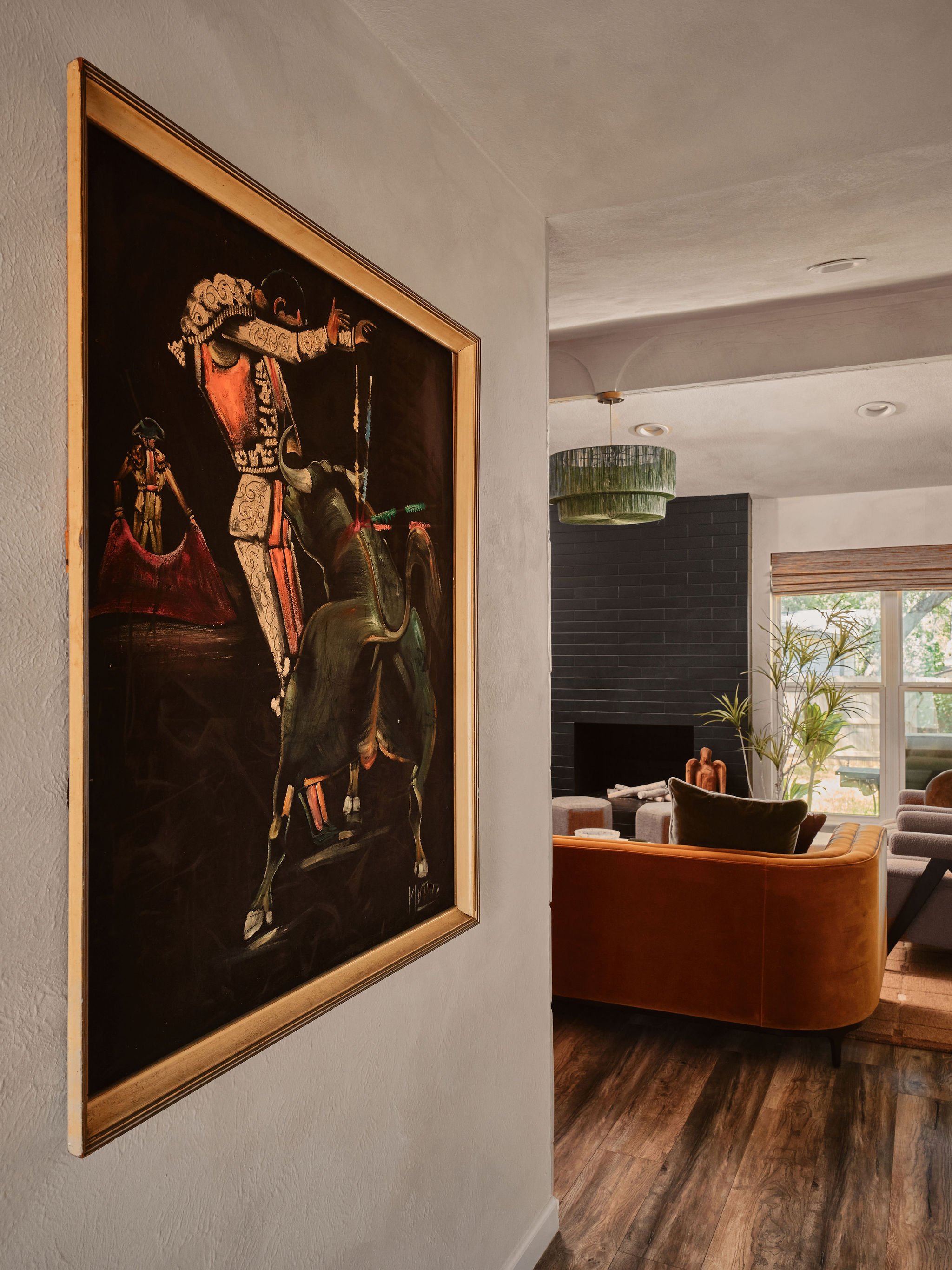 Living room with a large painting of a woman riding a horse, green hanging light fixture, black brick fireplace, window with blinds, and some plants.