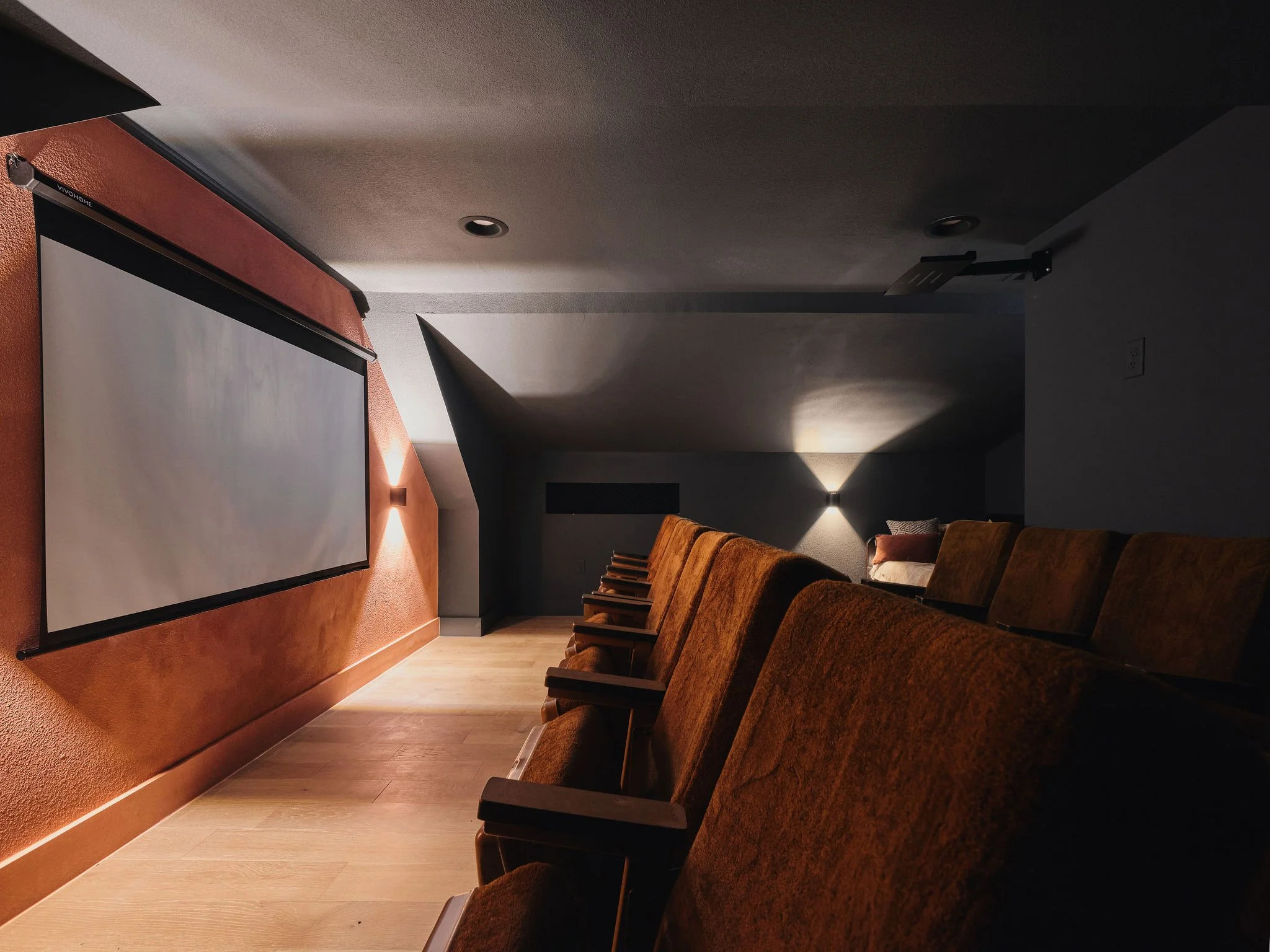 A dimly lit movie theater with plush brown seats arranged in rows, a large projection screen, and overhead lighting along the wall.