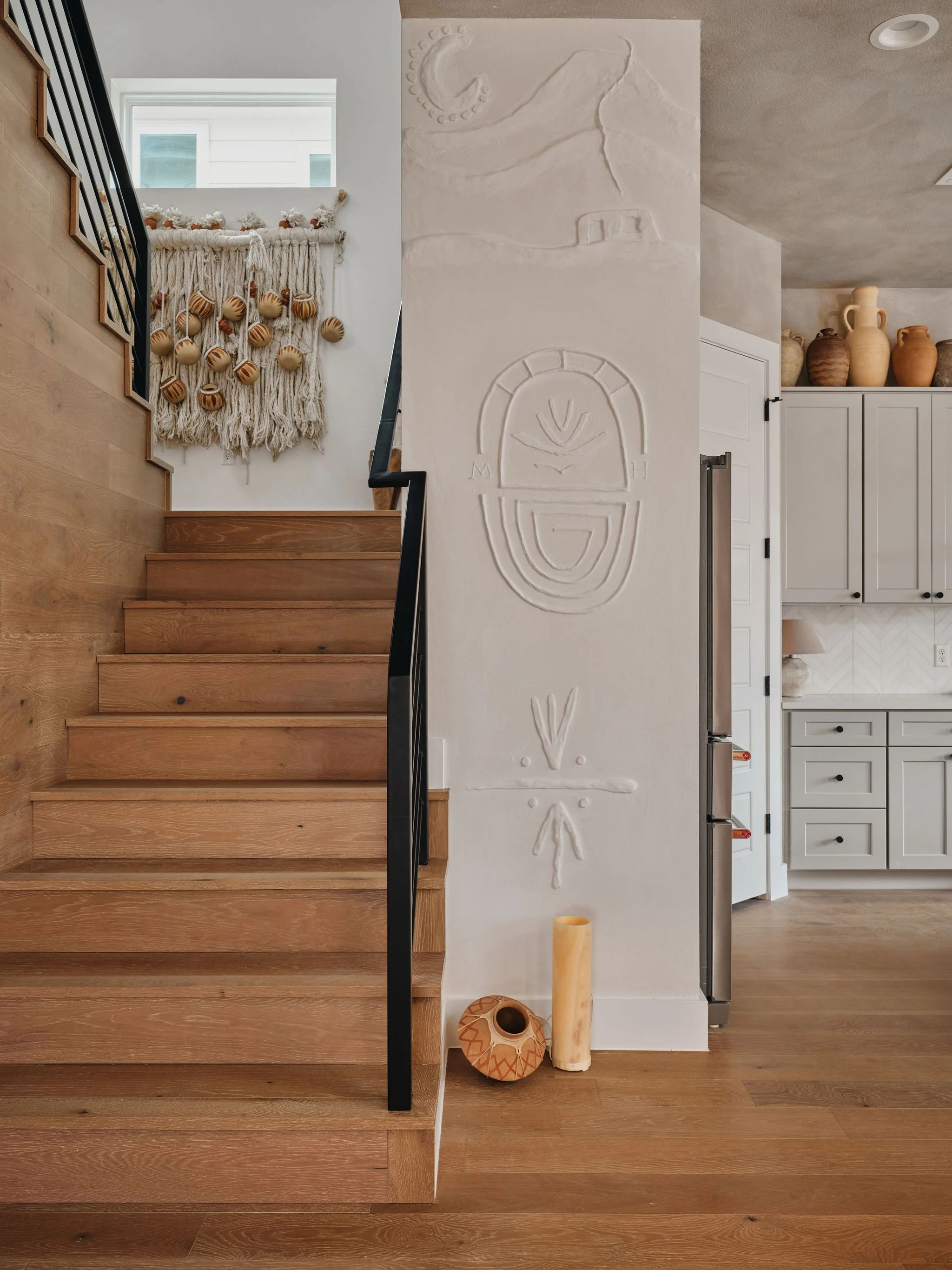 Interior view of a home showing wooden stairs, a decorative wall with primitive art, and a kitchen with white cabinetry and potted vases on top.