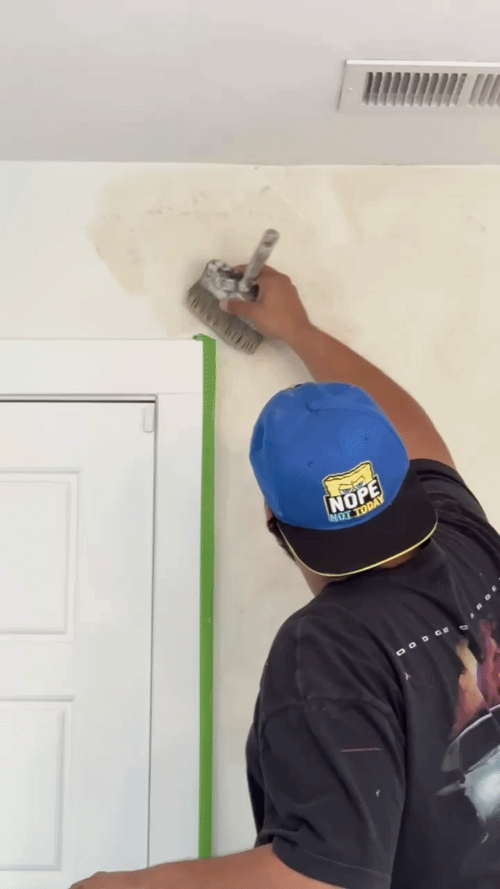 Person painting a wall near a white door frame using a paint roller, with green painter's tape on the edges.